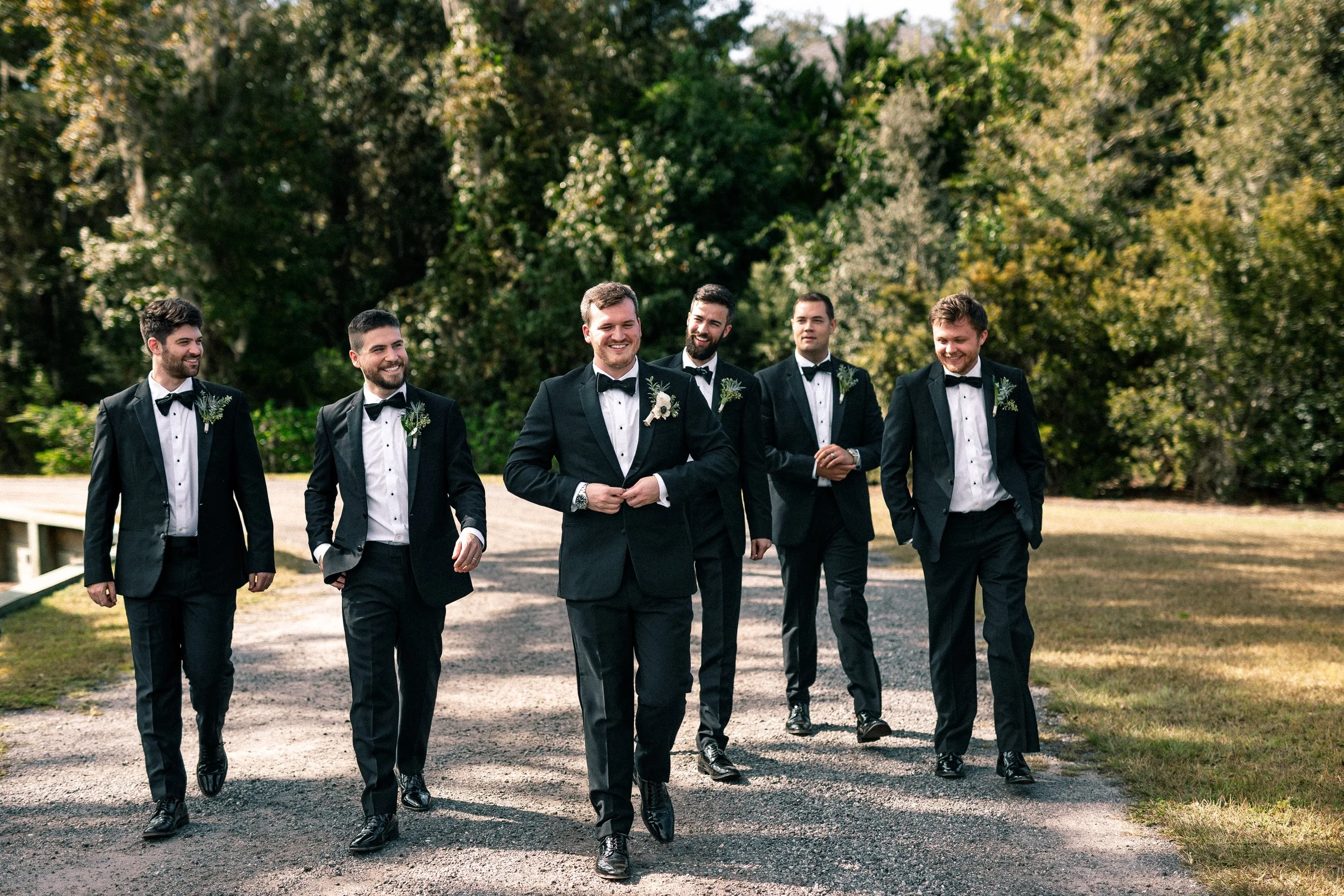 A group of six men in tuxedos walking on a gravel path in a park-like setting, smiling and enjoying each other's company on a sunny day.