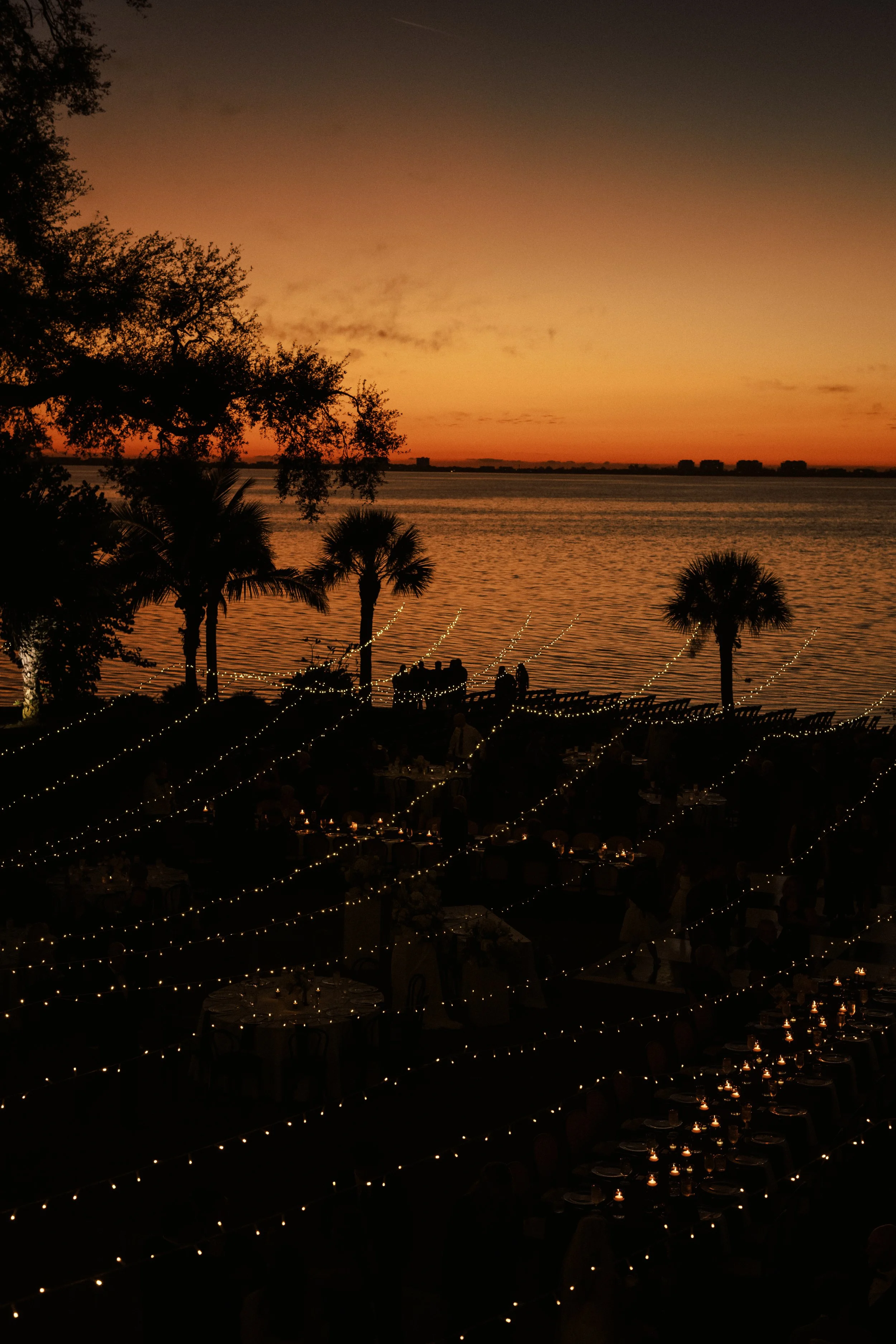 Sunset over a waterfront with trees and a decorated outdoor dining area with string lights and candles.