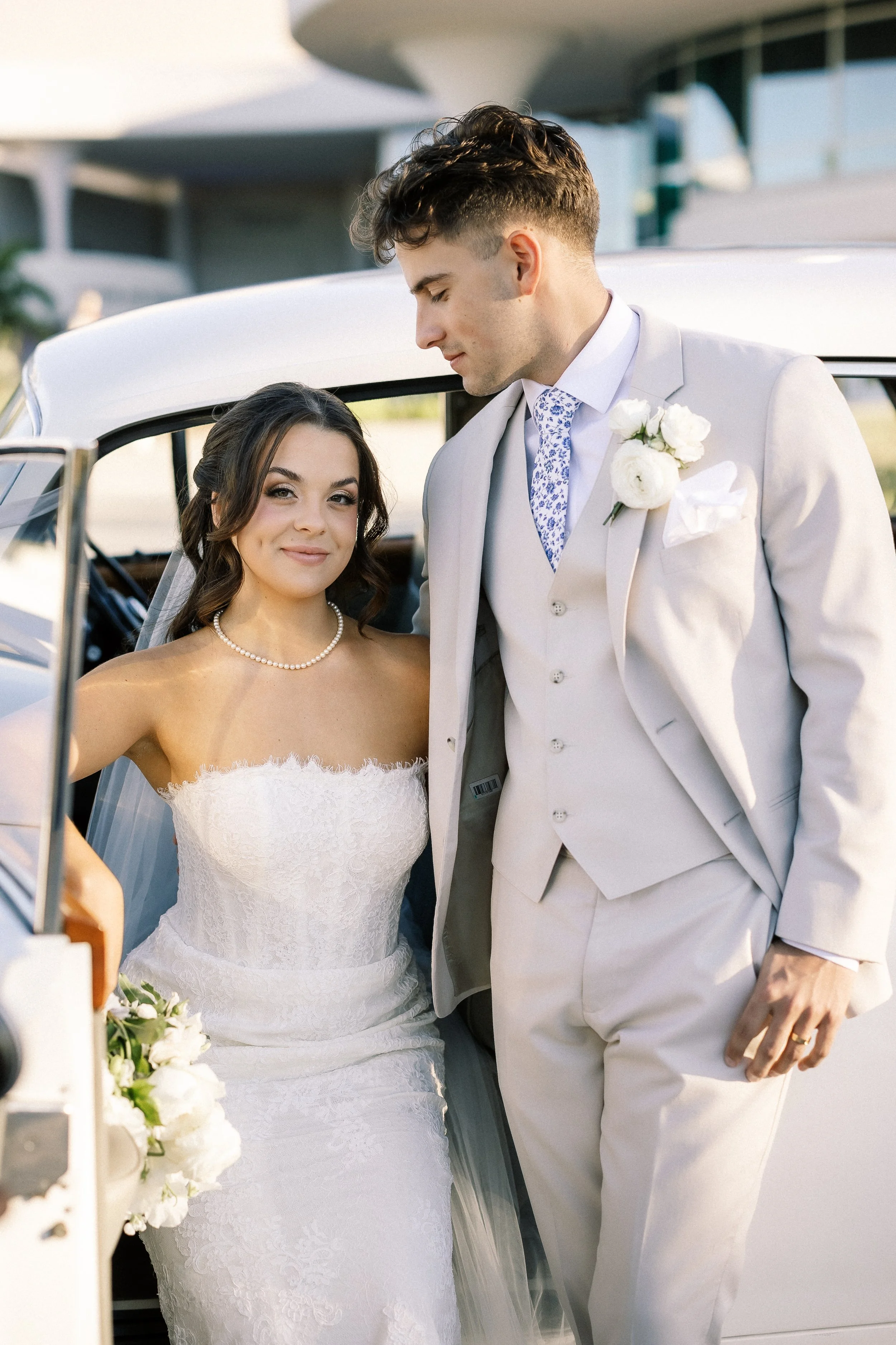 A bride and groom standing beside a white vintage car outside during daytime. The bride is wearing a strapless lace wedding dress and pearl necklace, holding a bouquet. The groom is in a light-colored suit with a flower boutonniere and a patterned ti