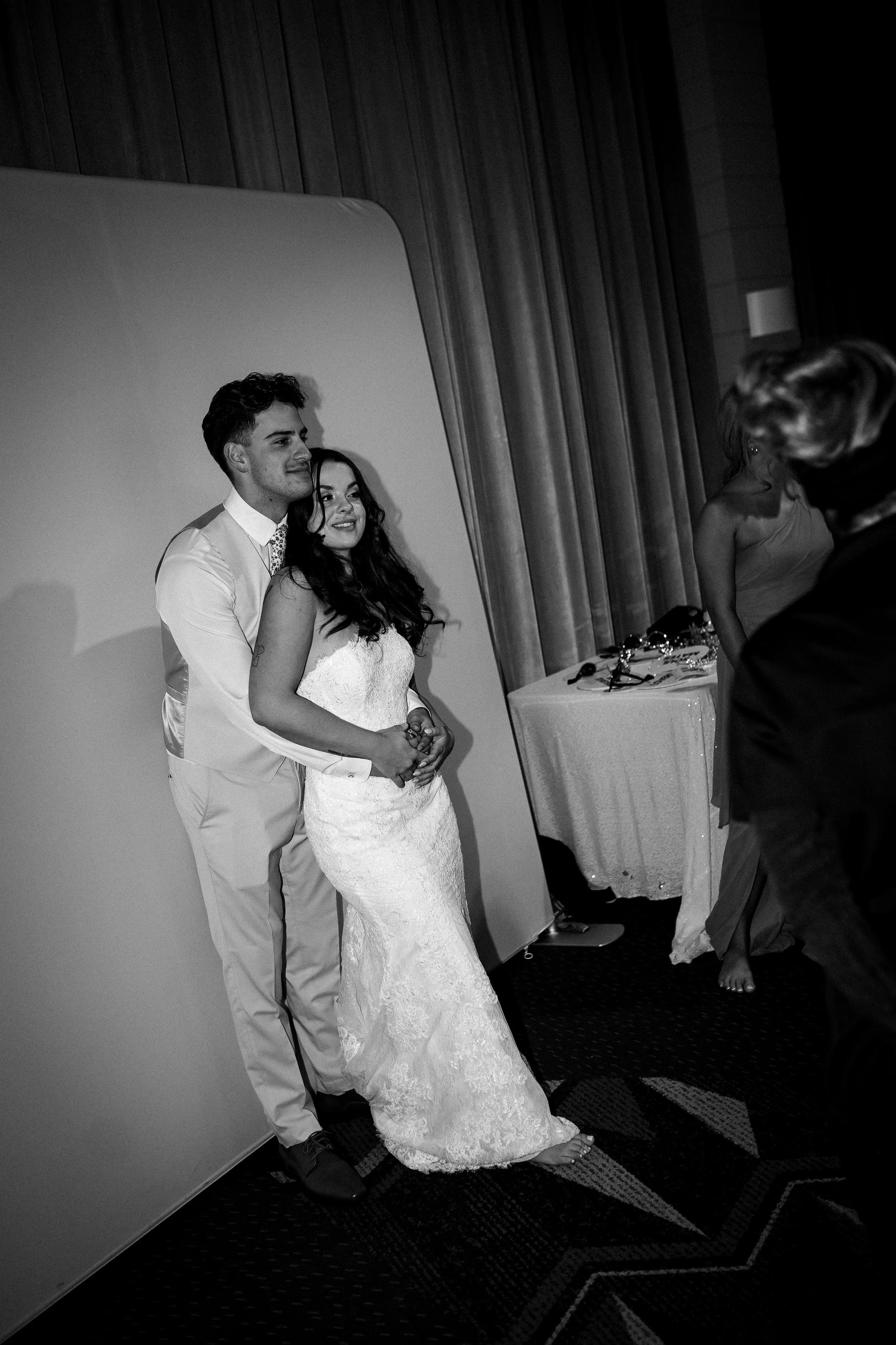 A bride and groom posing for a photo at their wedding reception, standing close together, with the groom hugging the bride from behind. The scene is in black and white, with a curtain and a table with some items visible in the background.