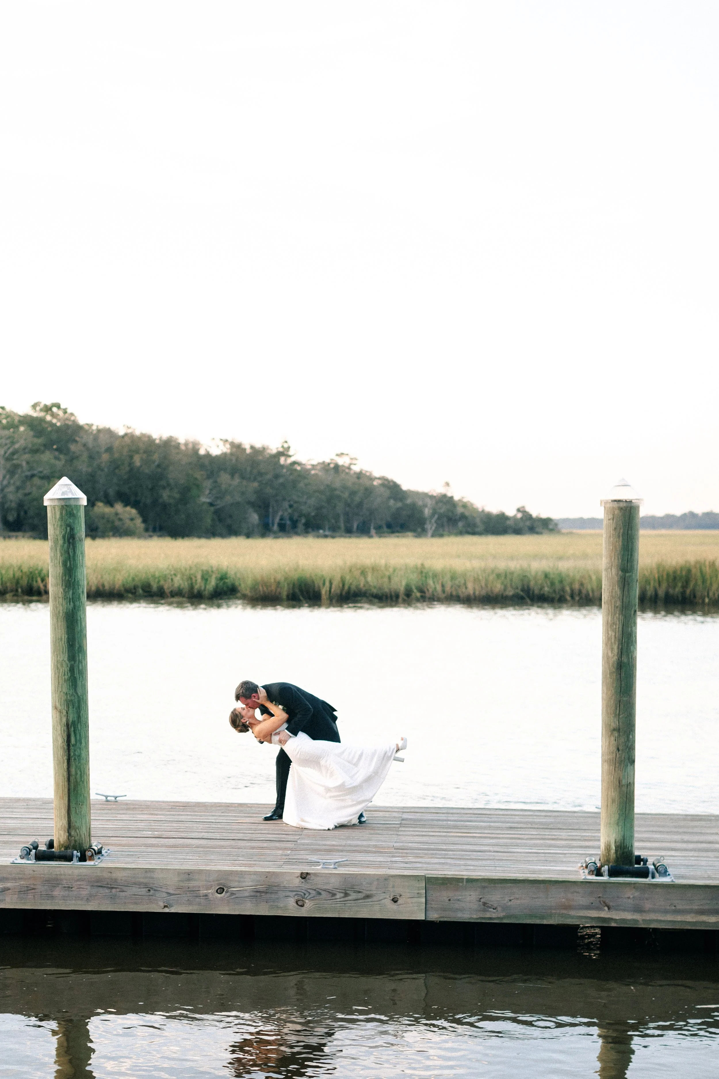 A couple in wedding attire sharing a kiss on a dock by the water, with a lush green landscape in the background.