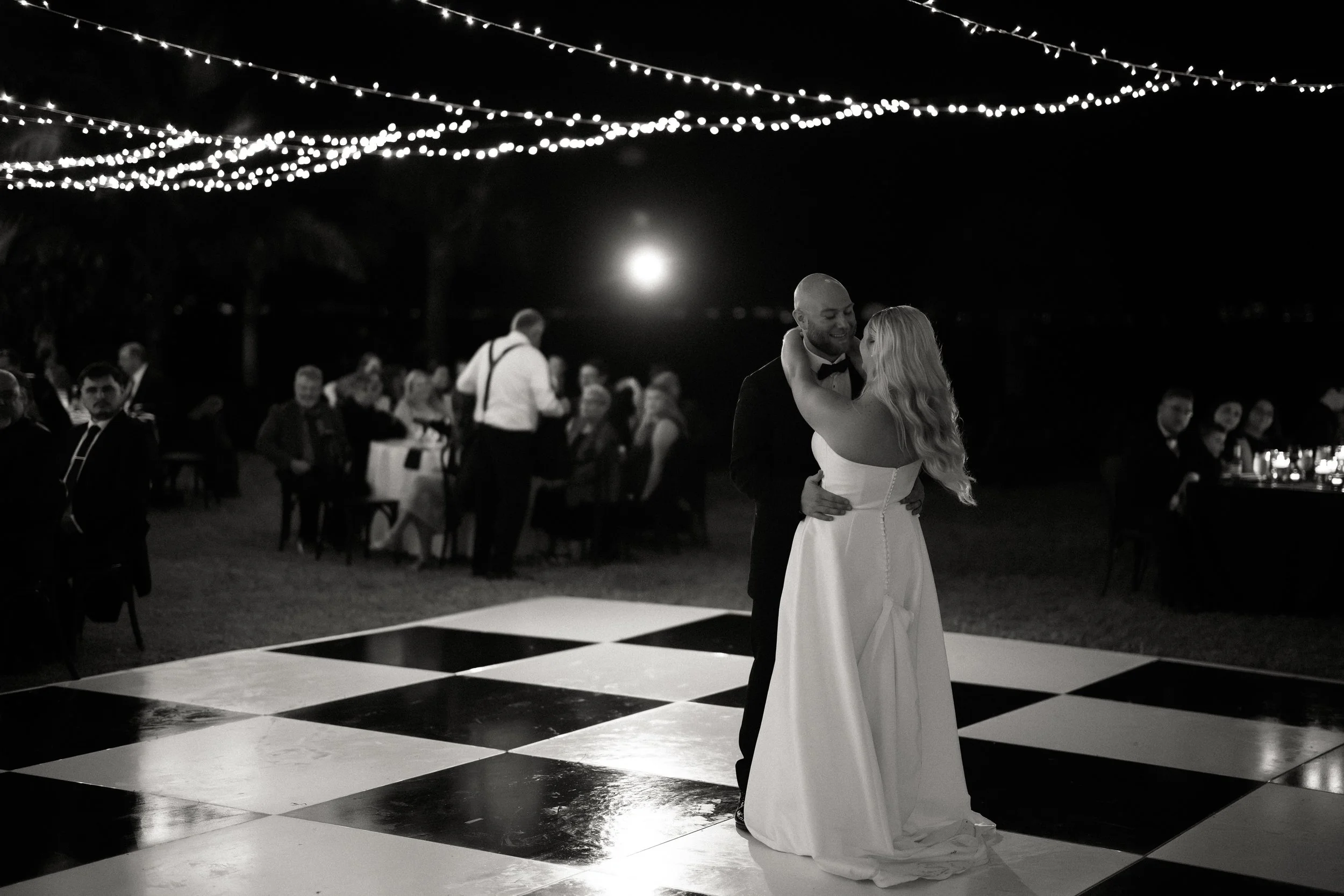 A bride and groom dancing on a checkered floor at night during a wedding reception, with guests seated at tables in the background and string lights overhead.