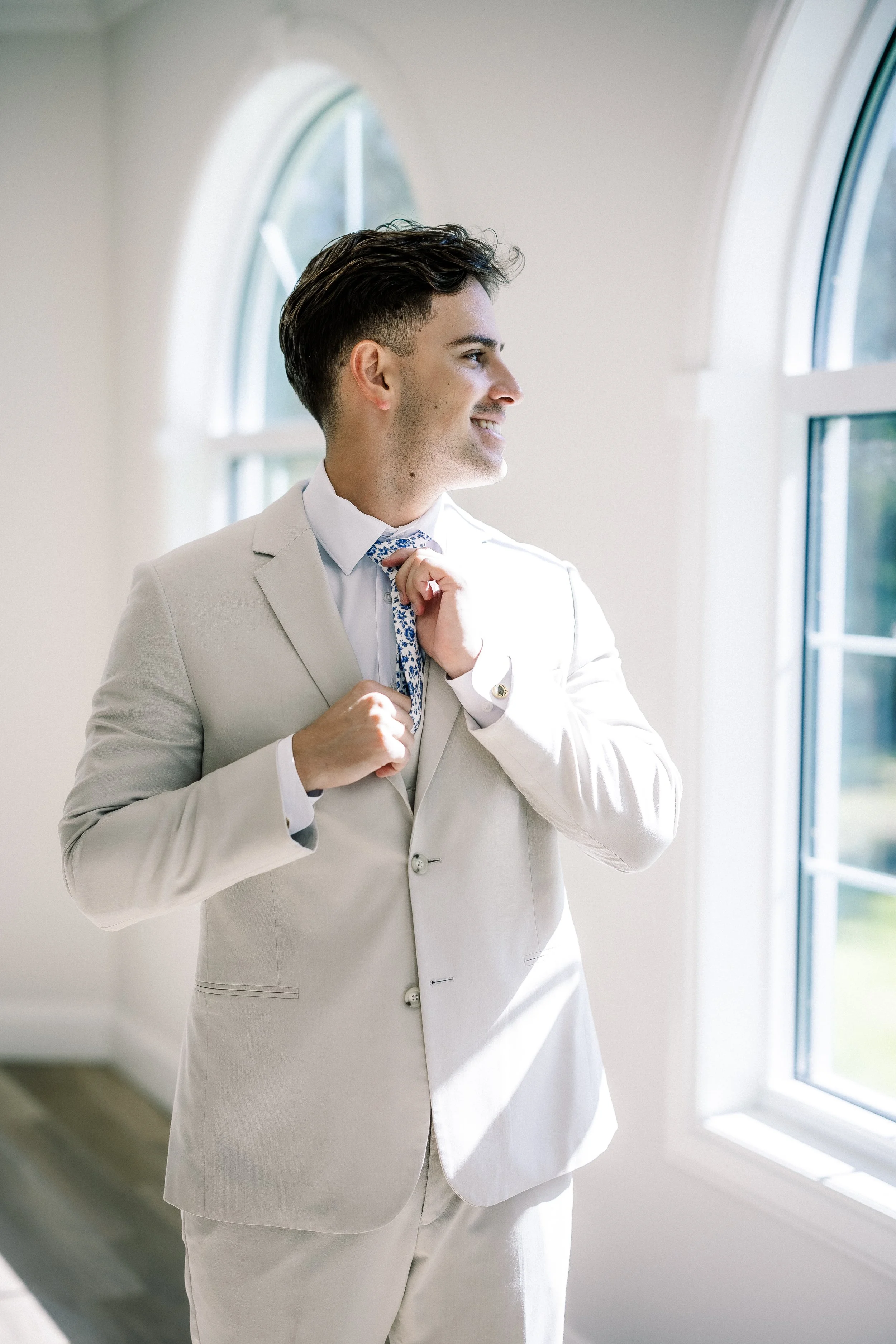 Young man in light beige suit adjusting his patterned tie while smiling, standing by a window with sunlight streaming in.