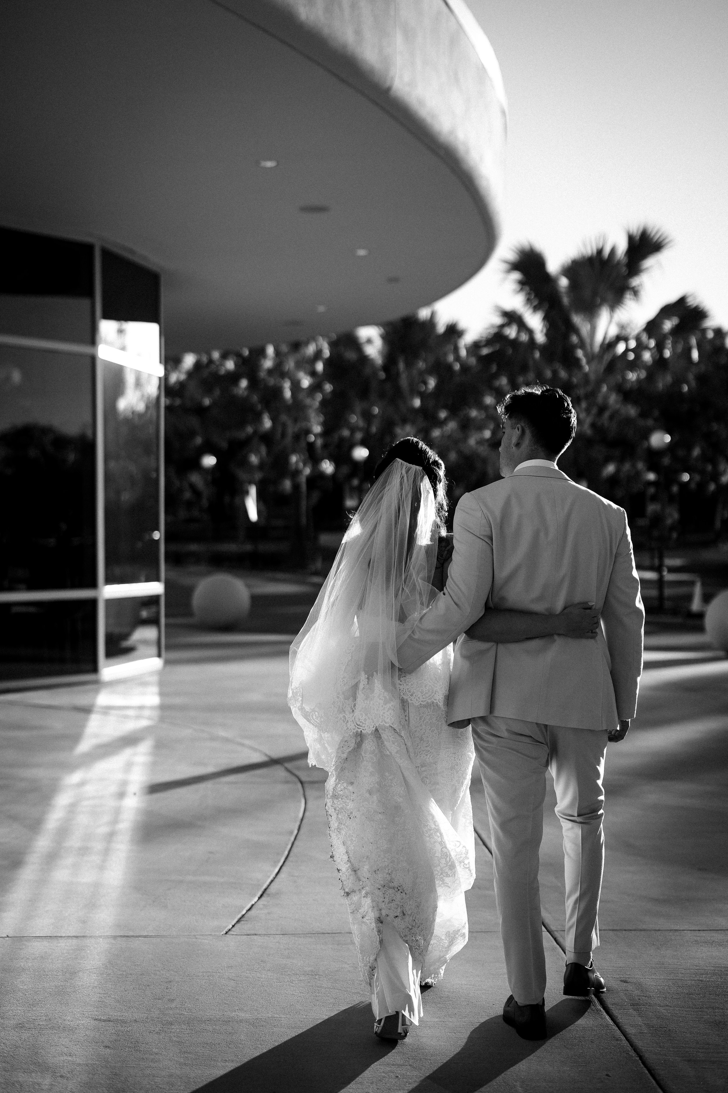 A black-and-white photo of a bride and groom walking arm-in-arm outdoors near a modern building and tropical trees during sunset.