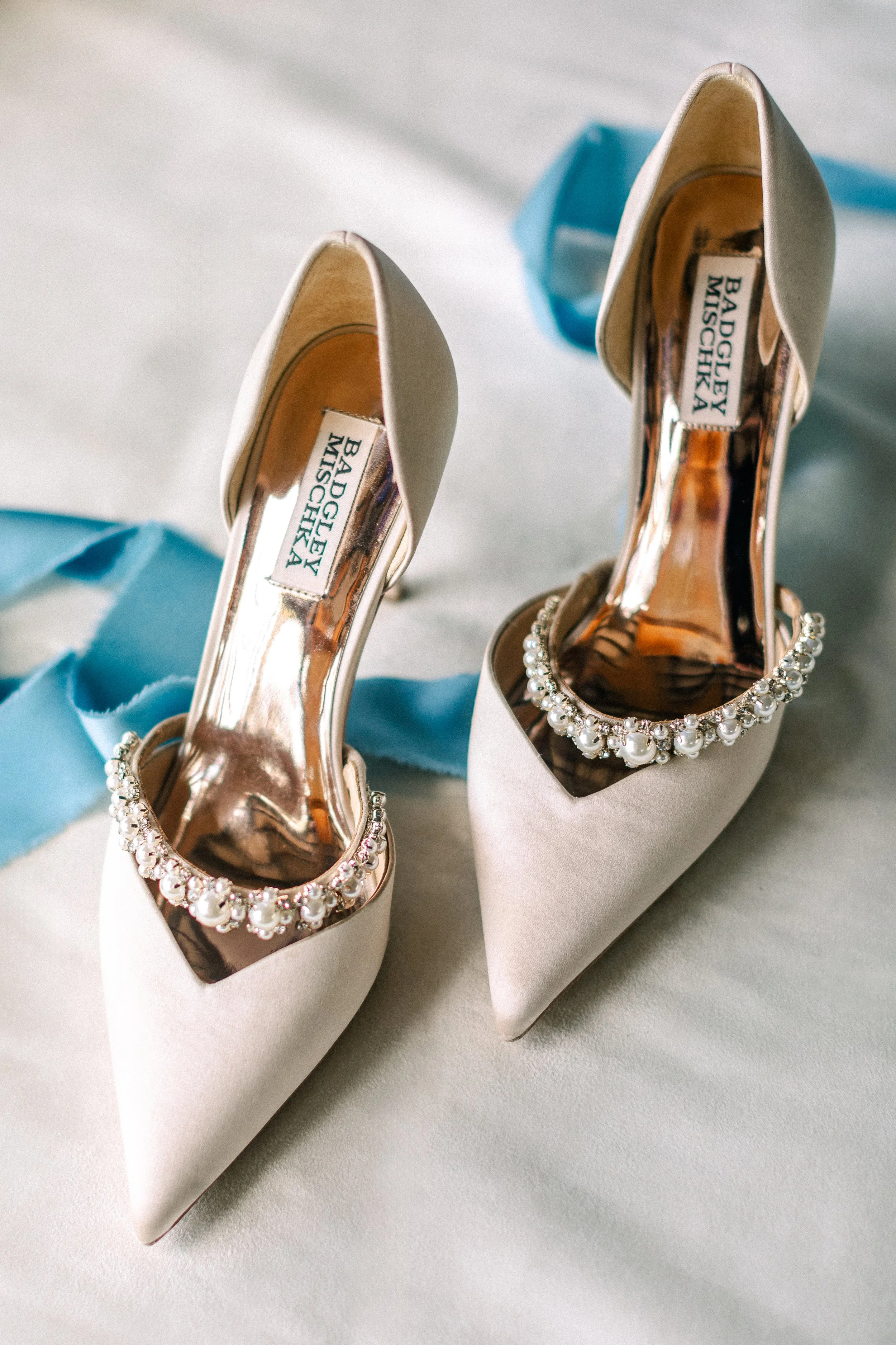 Pair of white high-heeled shoes adorned with pearls and gemstones, placed on a light-colored surface with light blue ribbons nearby.