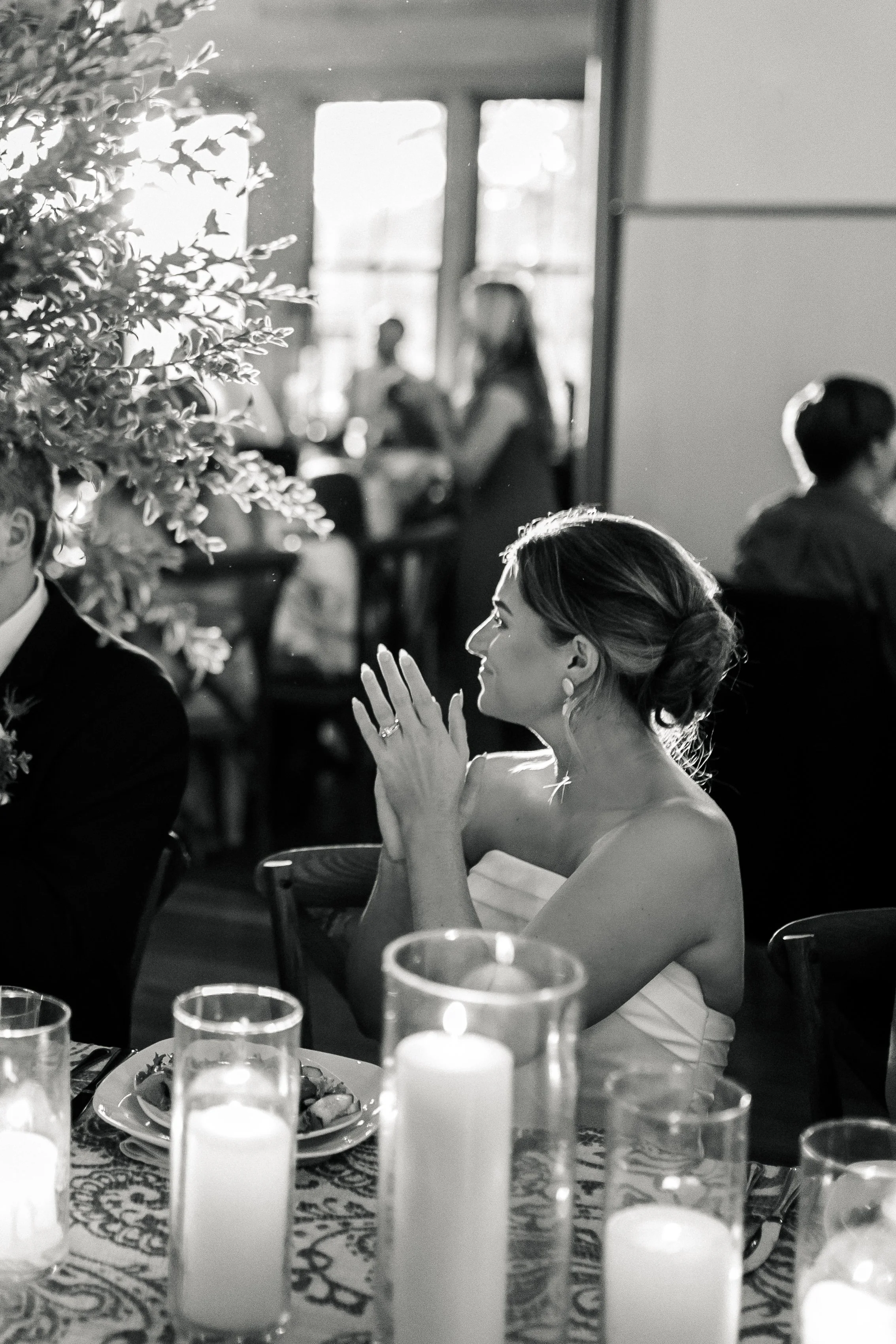 A woman in a strapless dress sitting at a table with lit candles, clapping her hands, during a formal gathering or celebration in a well-lit room, possibly a wedding reception.