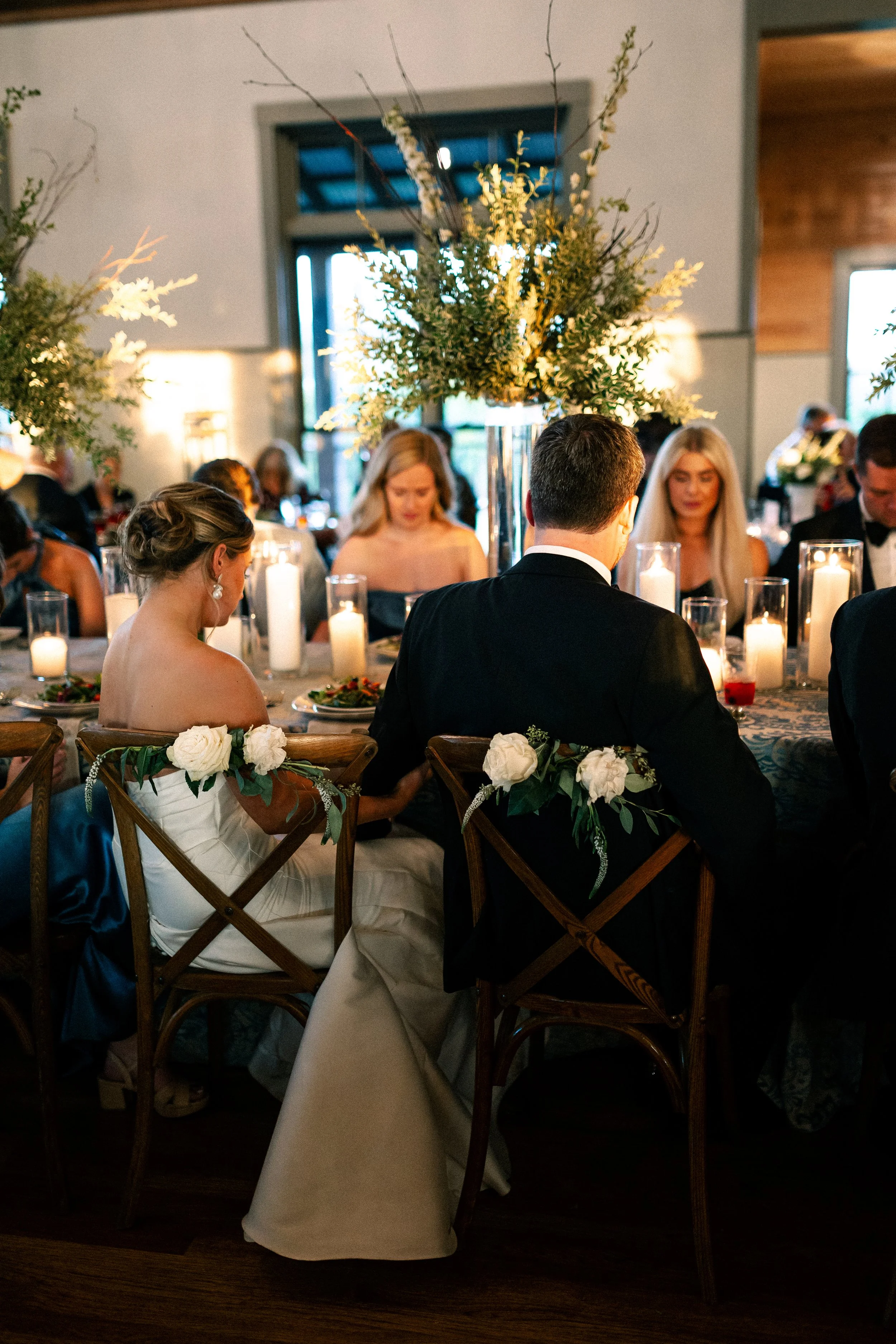 A wedding reception with a bride and groom seated at a decorated table, surrounded by guests, candles, and floral centerpieces.