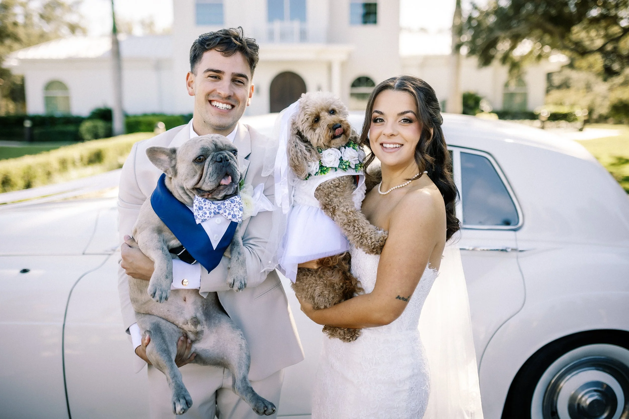 A smiling couple holding dogs in front of a vintage white car, with a house and trees in the background.