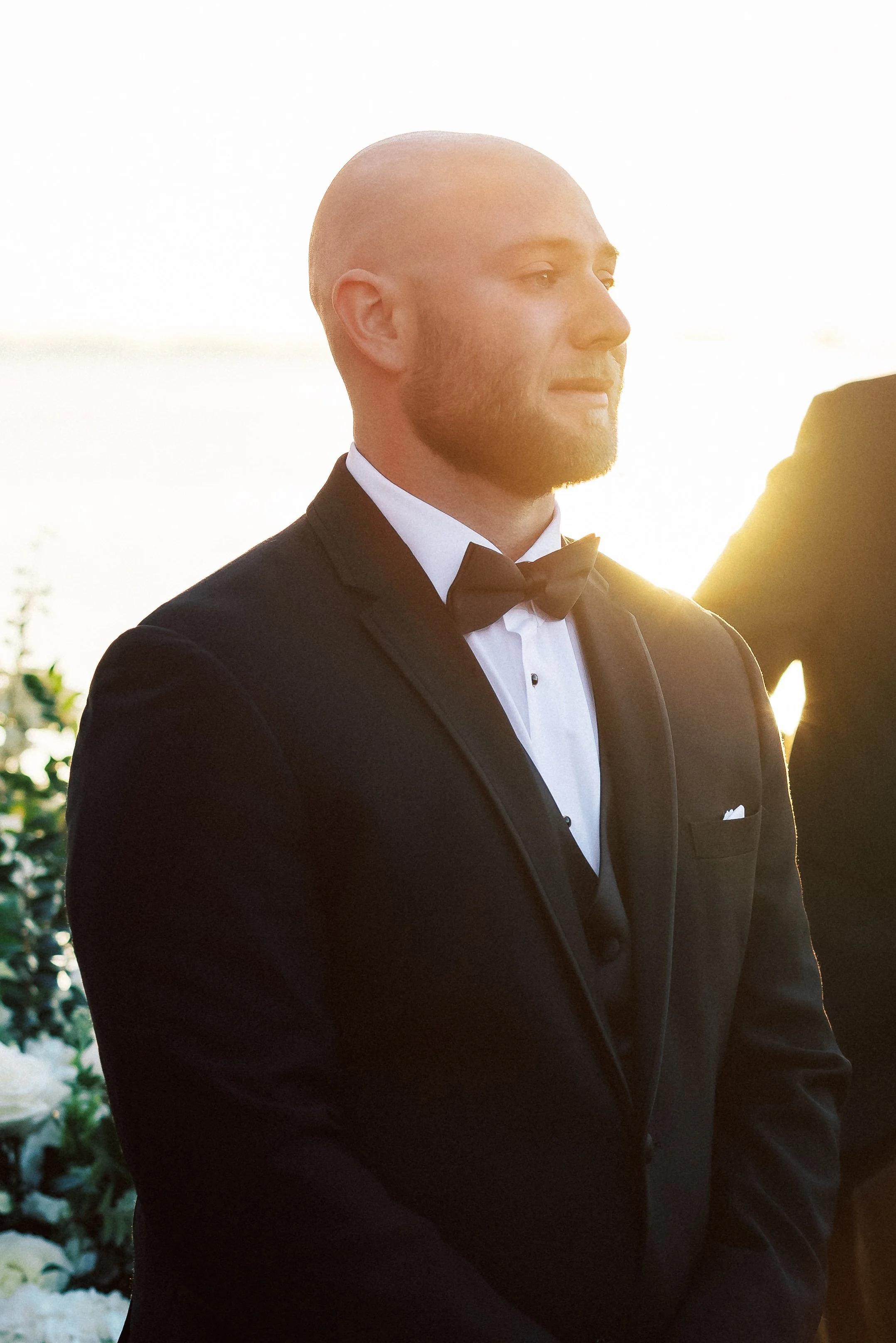A man in a black tuxedo with a bow tie, white shirt, and pocket square, standing outdoors at sunset.