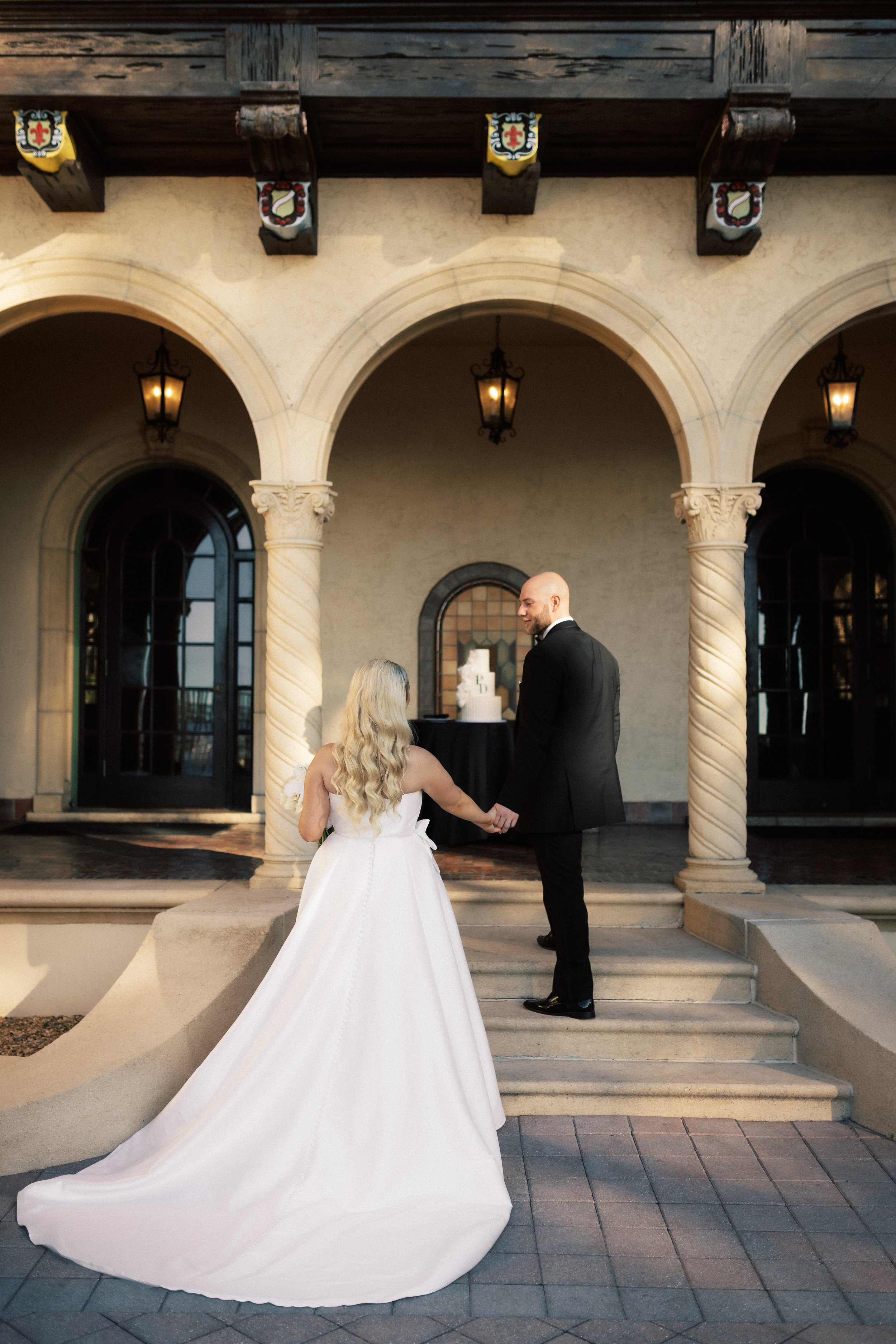 Bride and groom holding hands and exchanging vows on the steps of a building with arches, lanterns, and a wedding cake in the background.