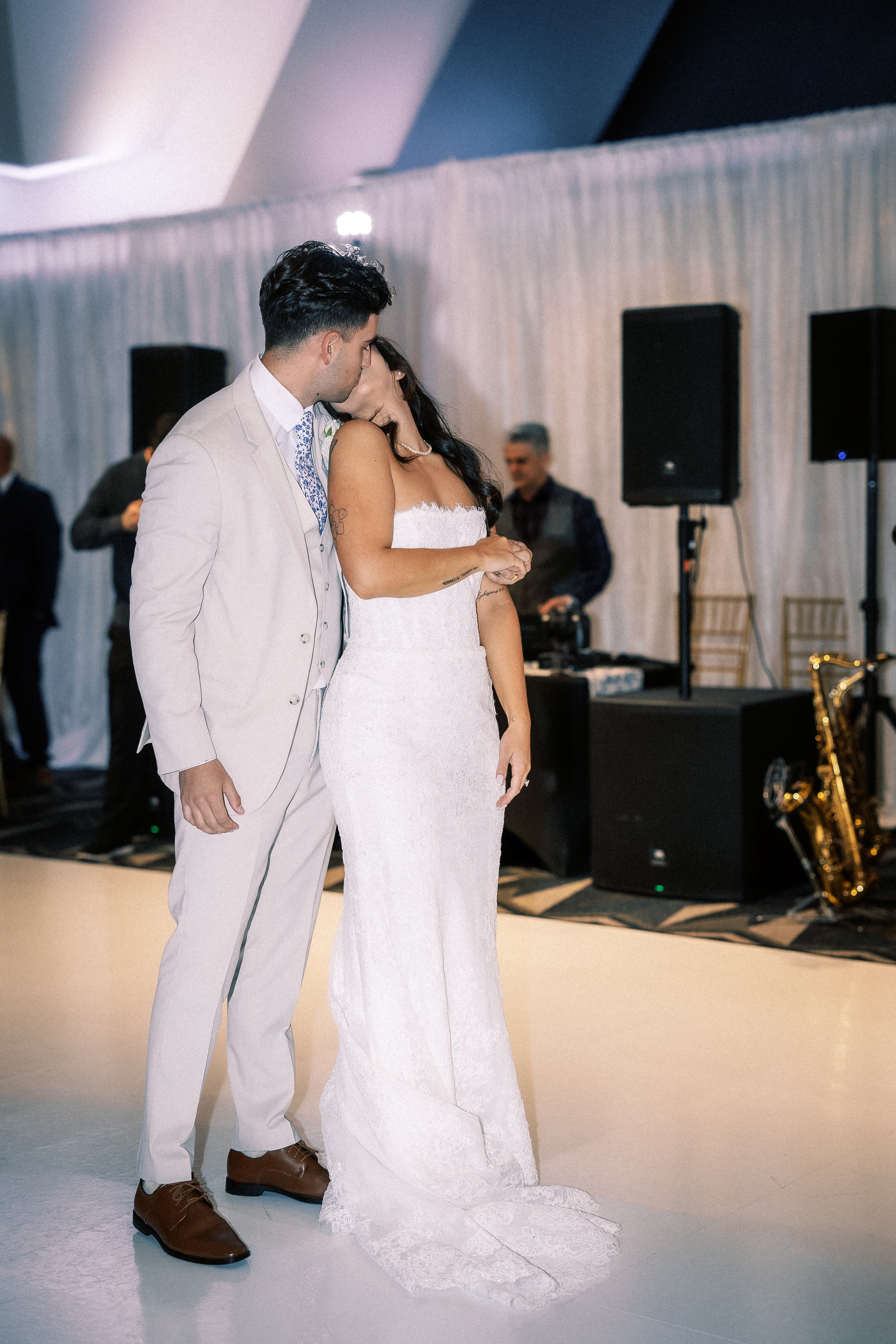 A bride and groom sharing a kiss during their wedding reception dance in a decorated venue with a DJ and speakers in the background.