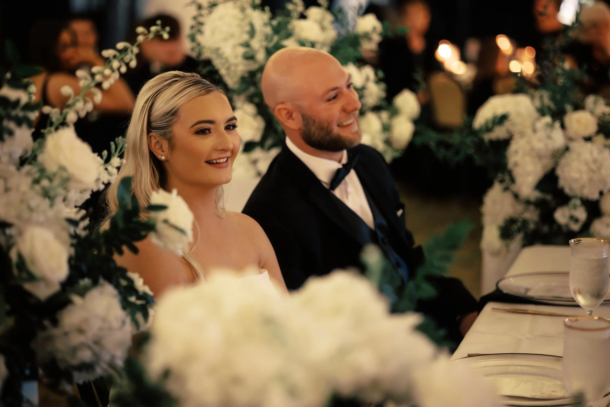 A smiling bride and groom sitting at a wedding reception table decorated with white flowers and greenery.