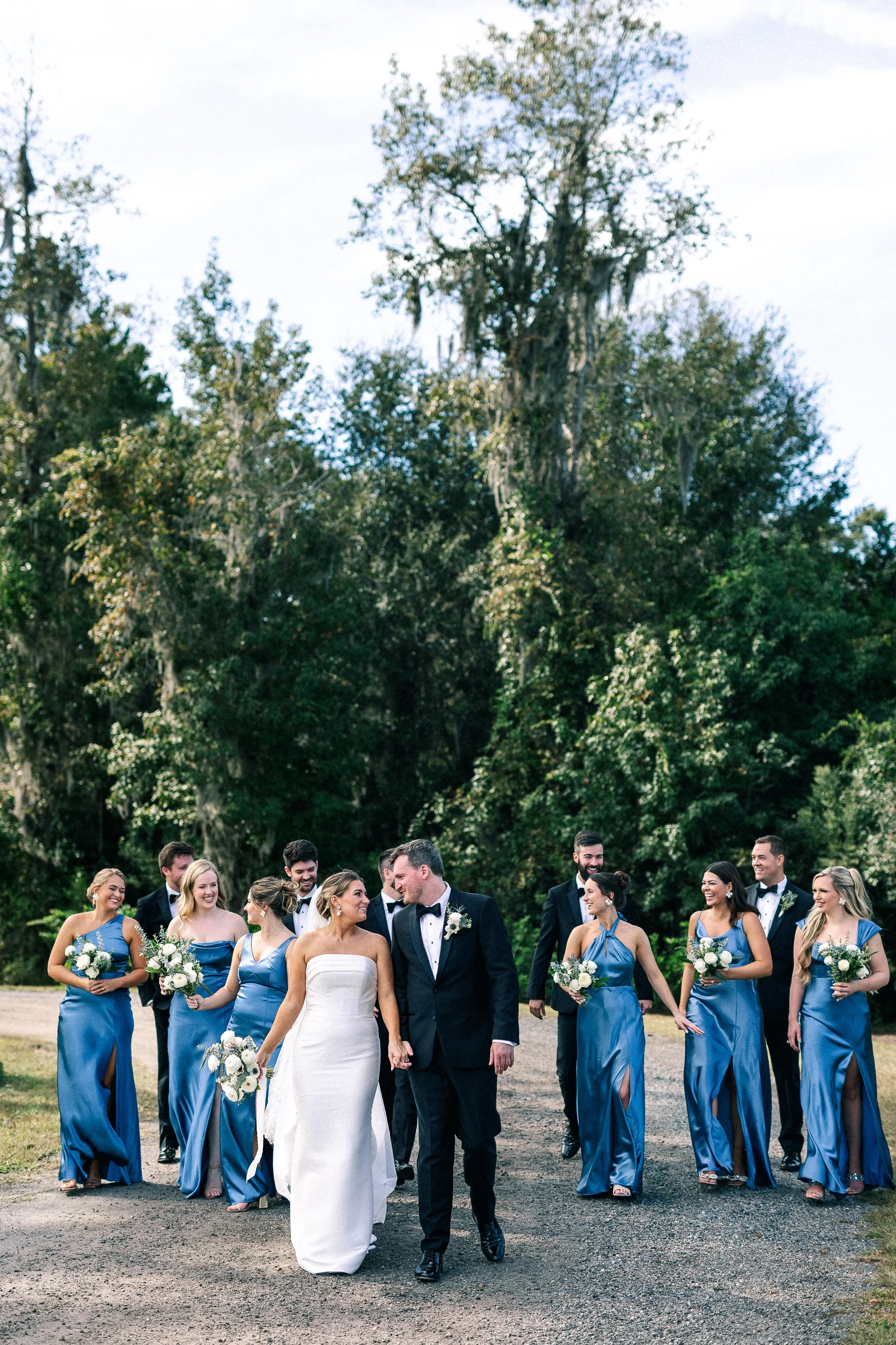 A wedding party walks outdoors on a gravel path, with the bride, groom, and bridesmaids in blue dresses, surrounded by tall trees.