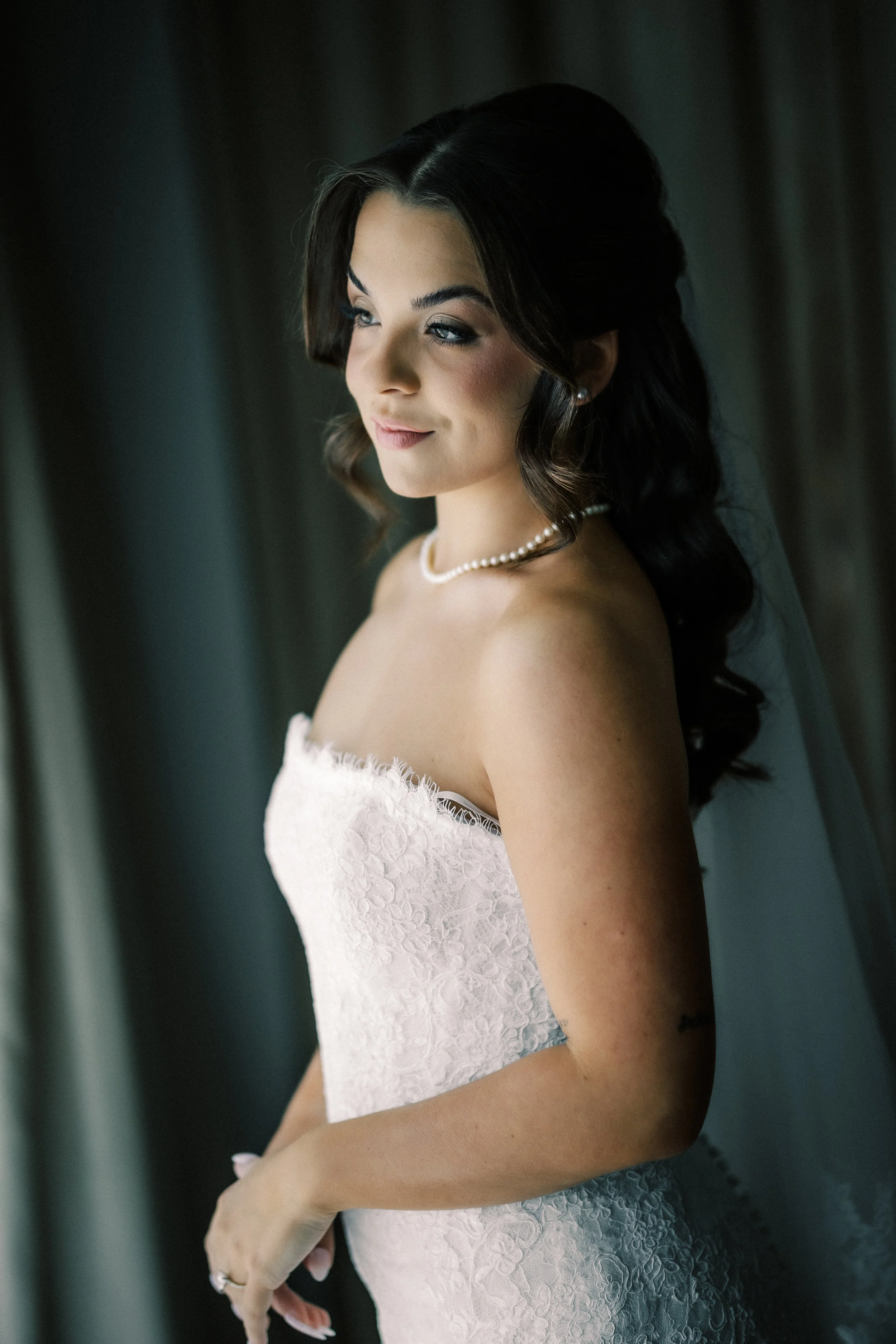 A bride with dark hair in loose curls, wearing a strapless white lace wedding dress, pearl necklace, and pearl earrings, standing near a window with soft natural light.