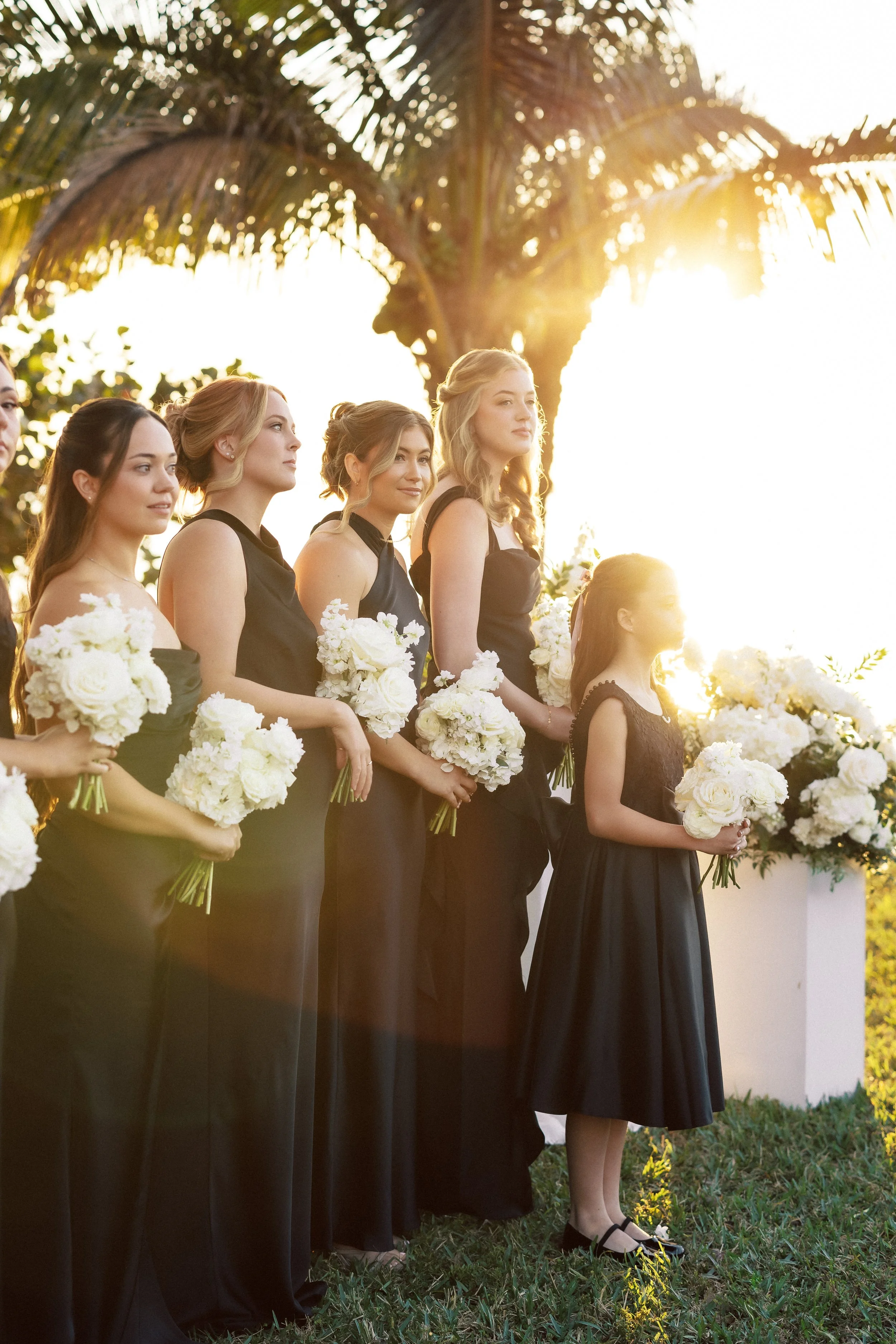 Group of bridesmaids and a young flower girl in black dresses holding white bouquets, standing outdoors under a palm tree with sunlight in the background.