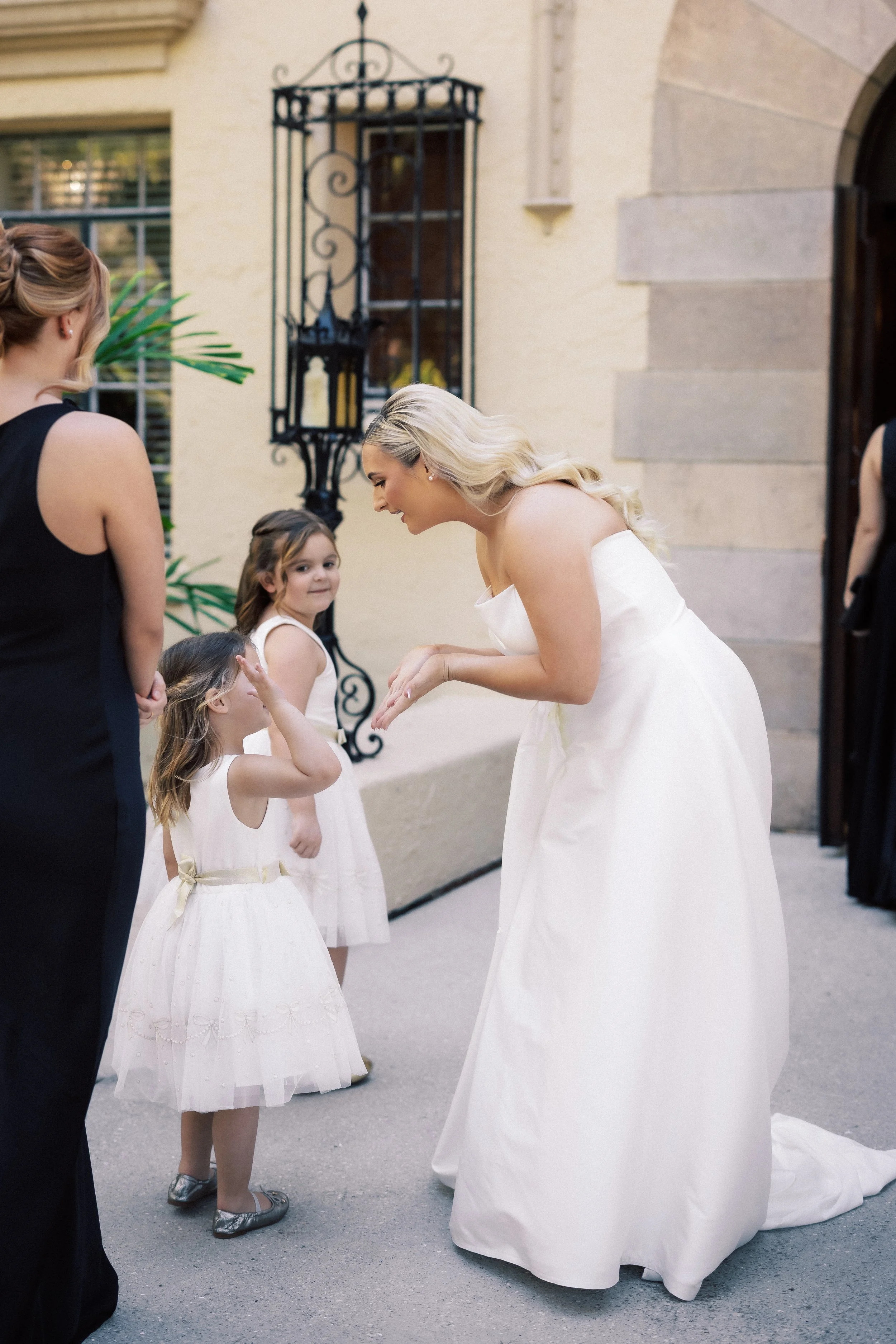A bride in a white wedding dress bends down to greet two young girls in white dresses at an outdoor wedding venue.