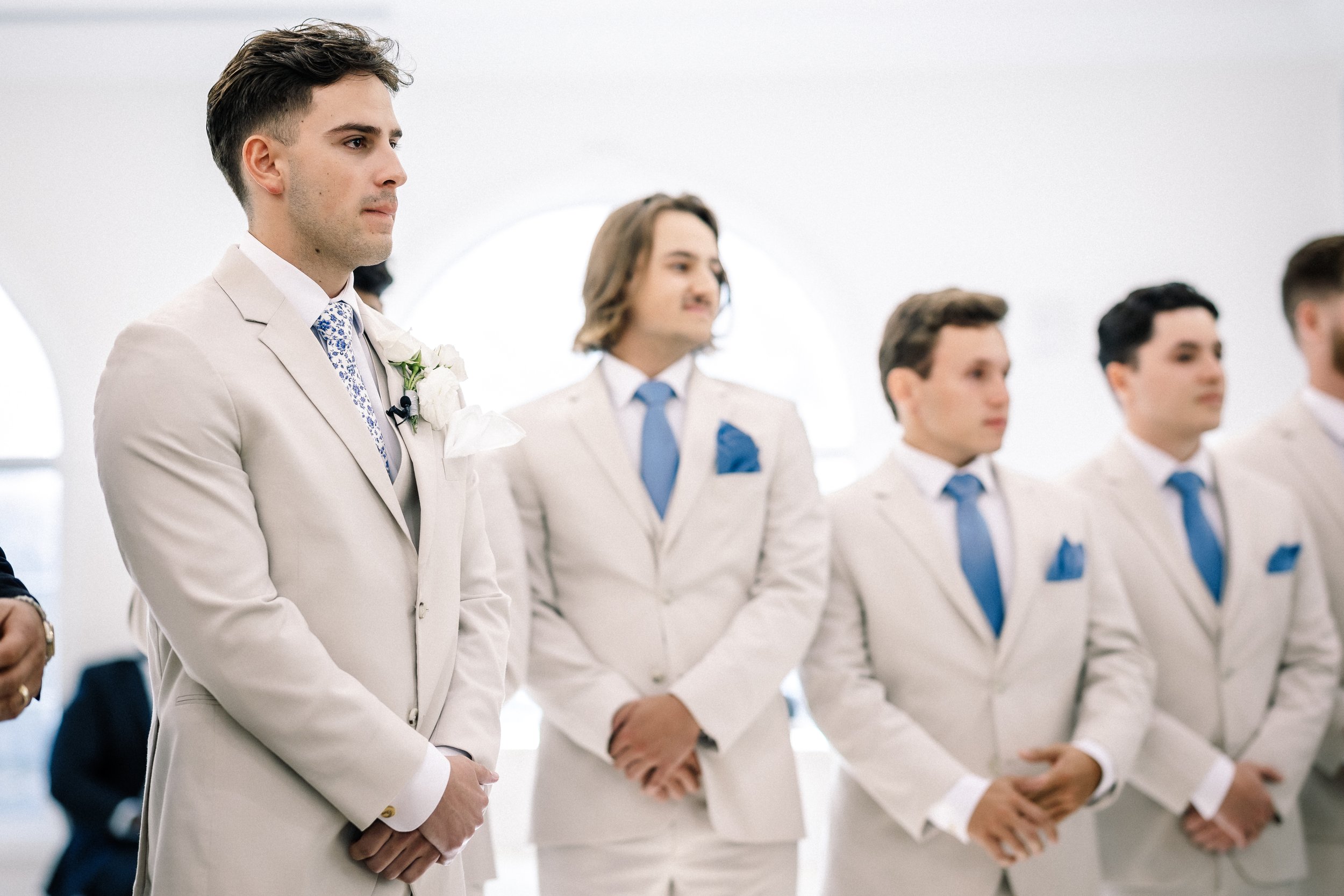 A group of men in white suits and blue ties standing in a line indoors during a formal event or wedding.