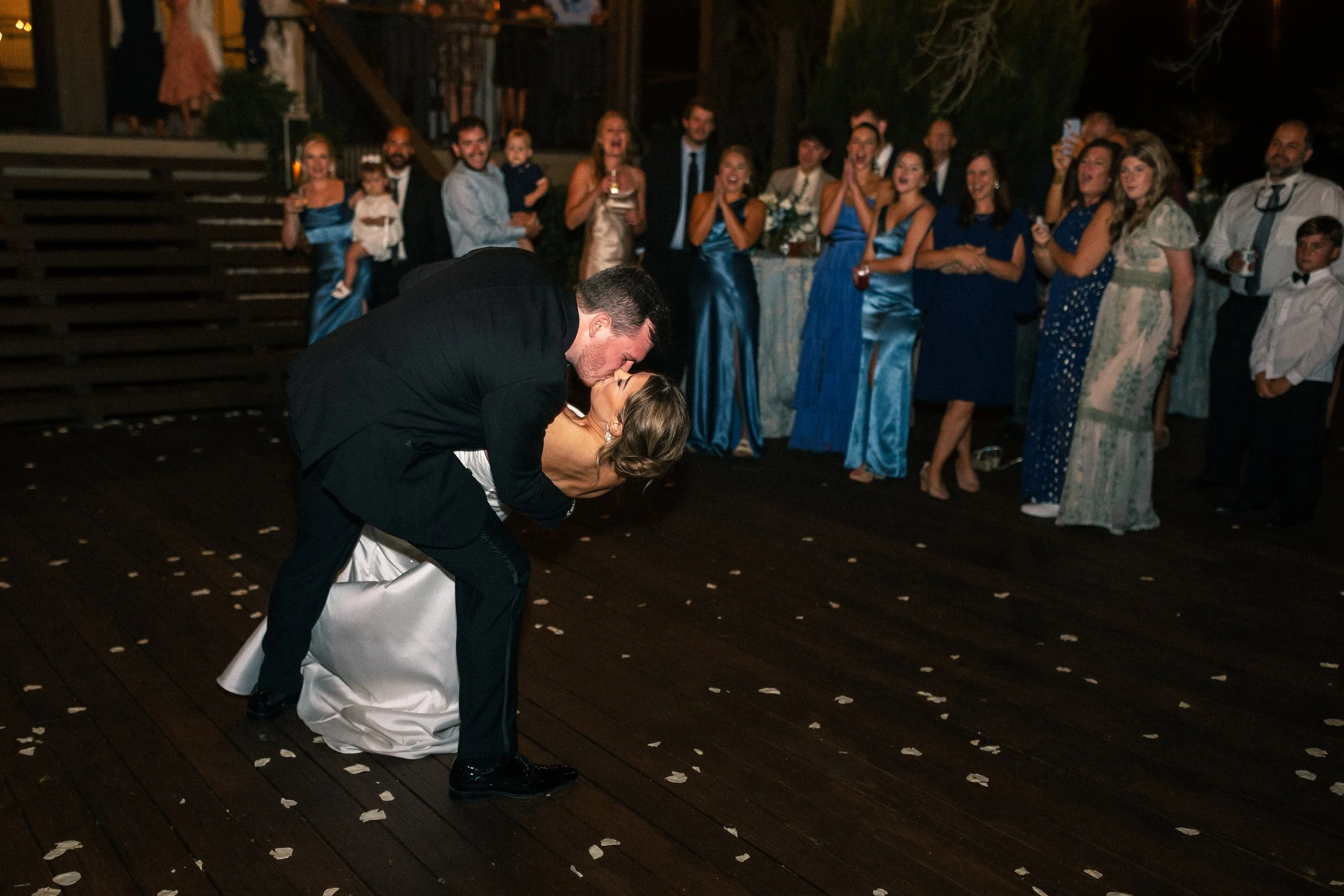 A bride and groom sharing a kiss during their wedding dance, with wedding guests watching and celebrating in the background.