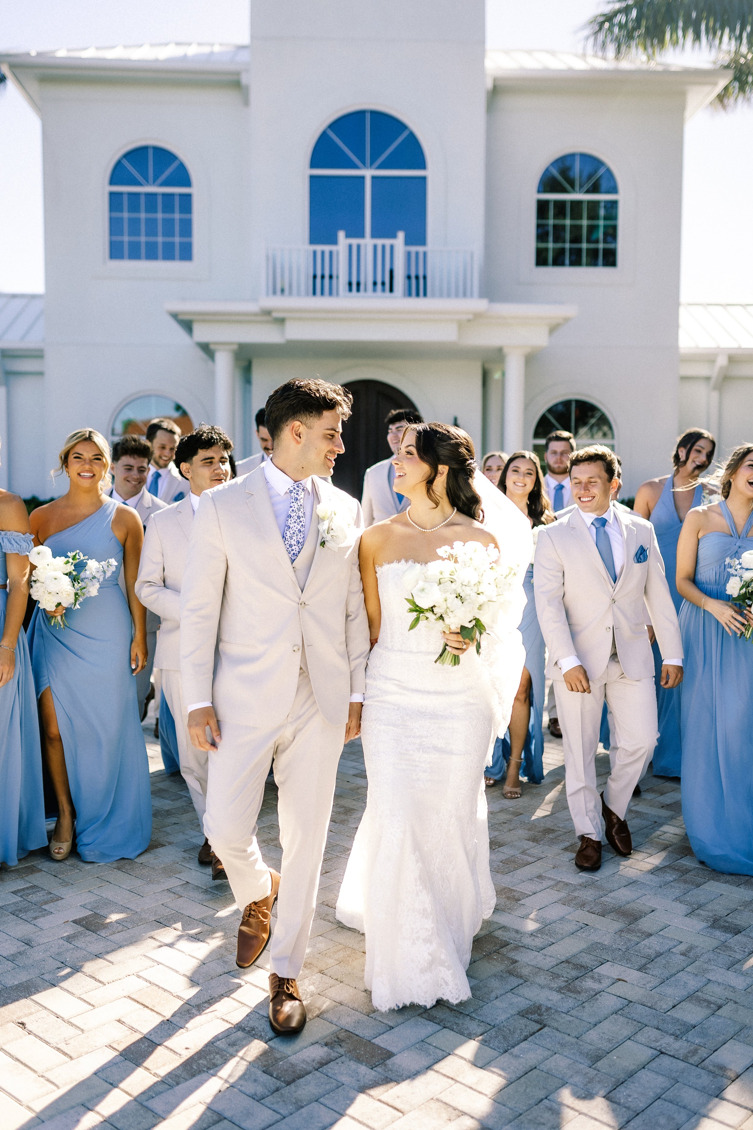 A bride and groom walk hand in hand outside a white house with large windows, surrounded by bridesmaids and groomsmen. The bride wears a white lace wedding dress and holds a bouquet of white flowers. The groom wears a light-colored suit with a patter