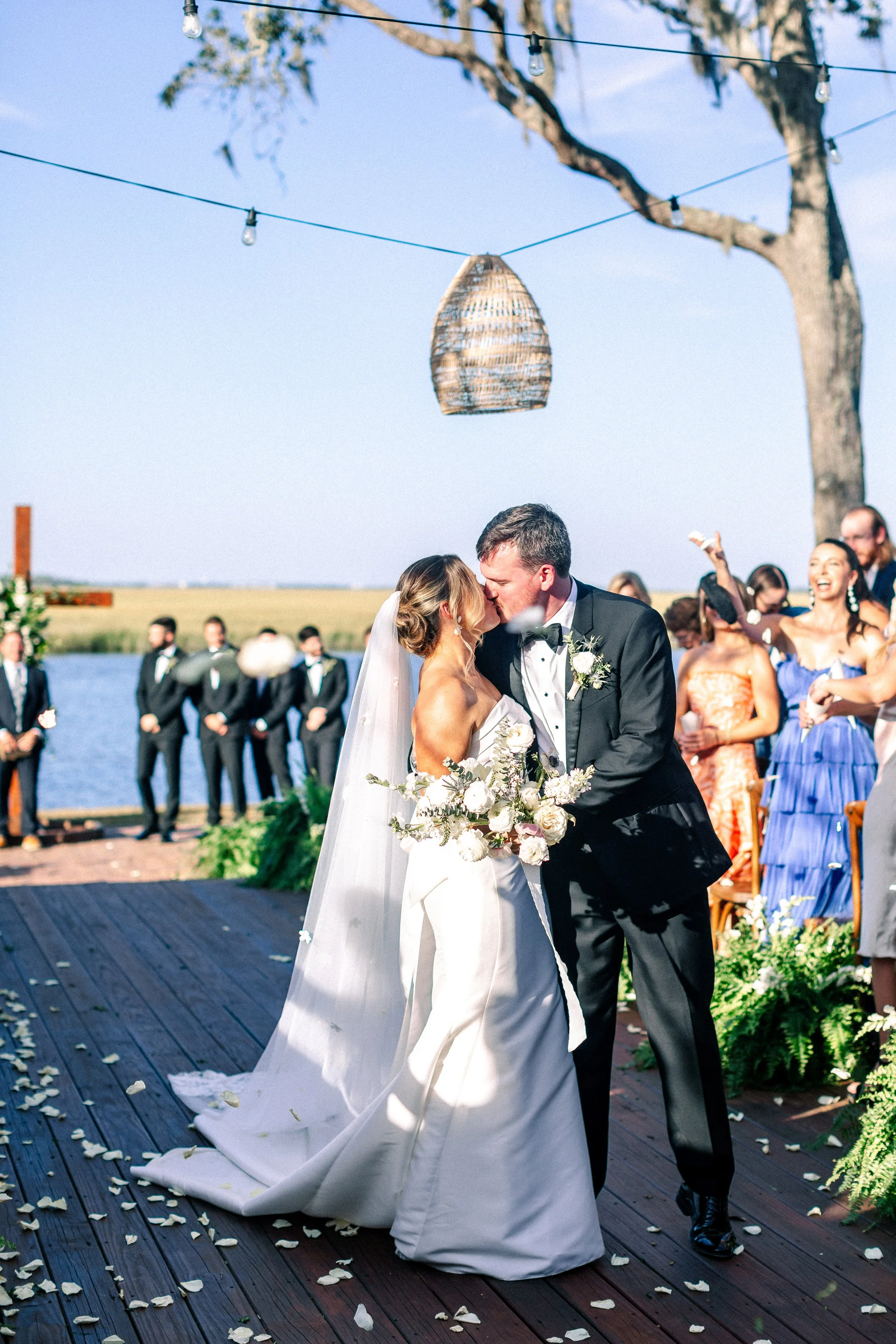 A bride and groom kissing during their outdoor wedding ceremony by a lake, with guests and bridesmaids in colorful dresses watching and celebrating.