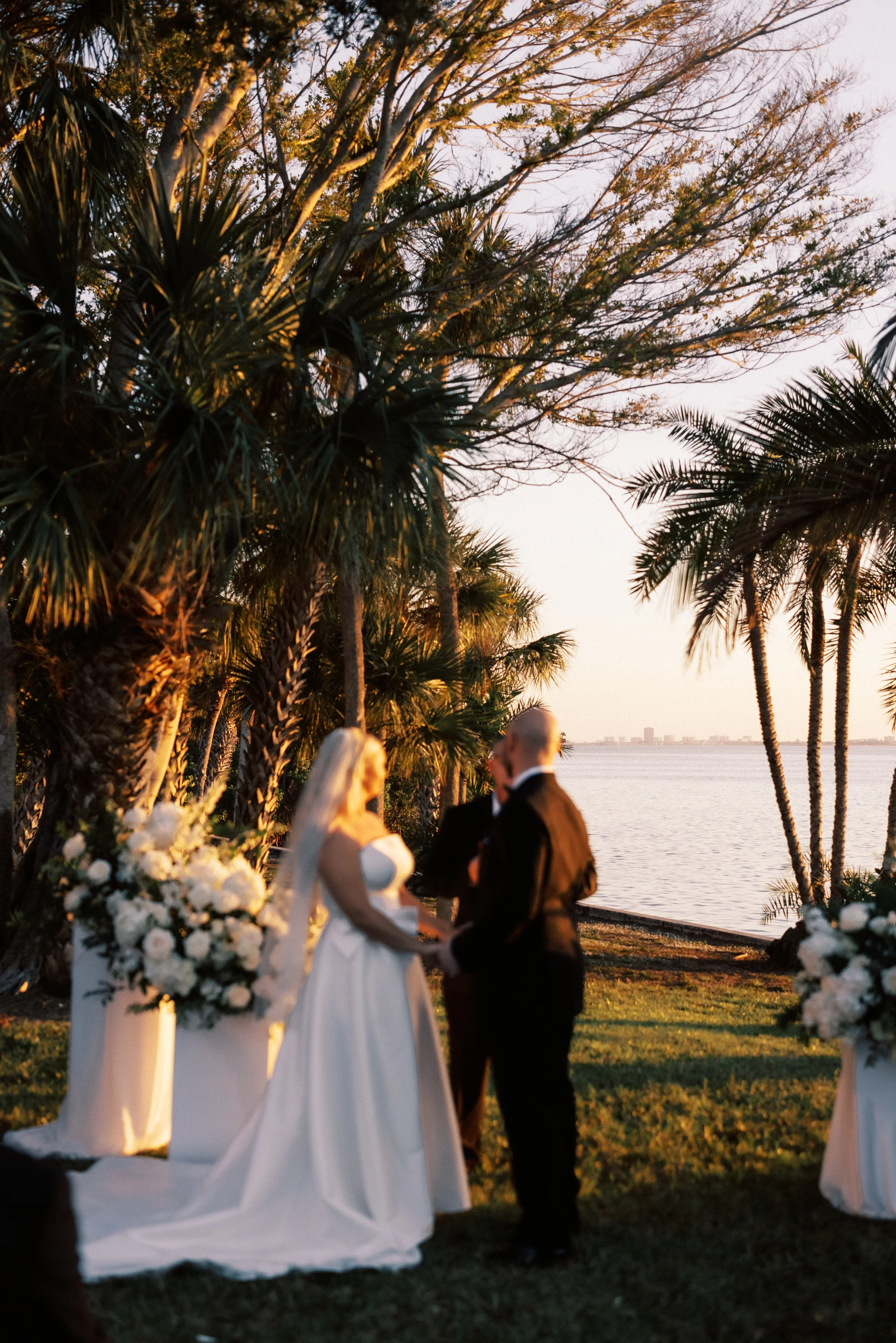 A wedding ceremony taking place outdoors near the water during sunset, with a bride and groom holding hands and facing each other, surrounded by trees and floral arrangements.