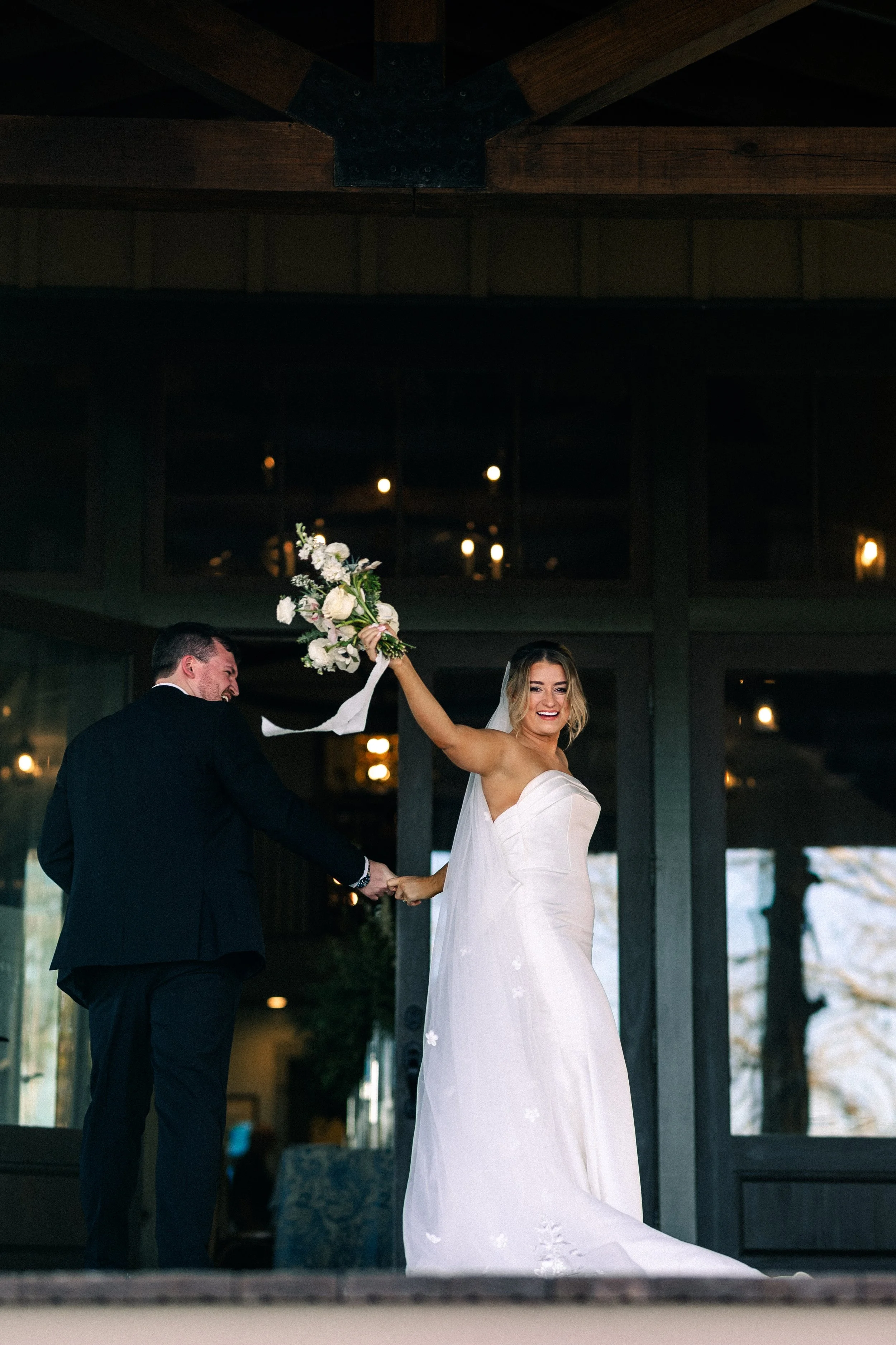 Bride in a white wedding dress holding a bouquet of flowers, smiling, while standing next to a groom in a black suit, indoor wedding venue with large windows and ambient lighting in the background.