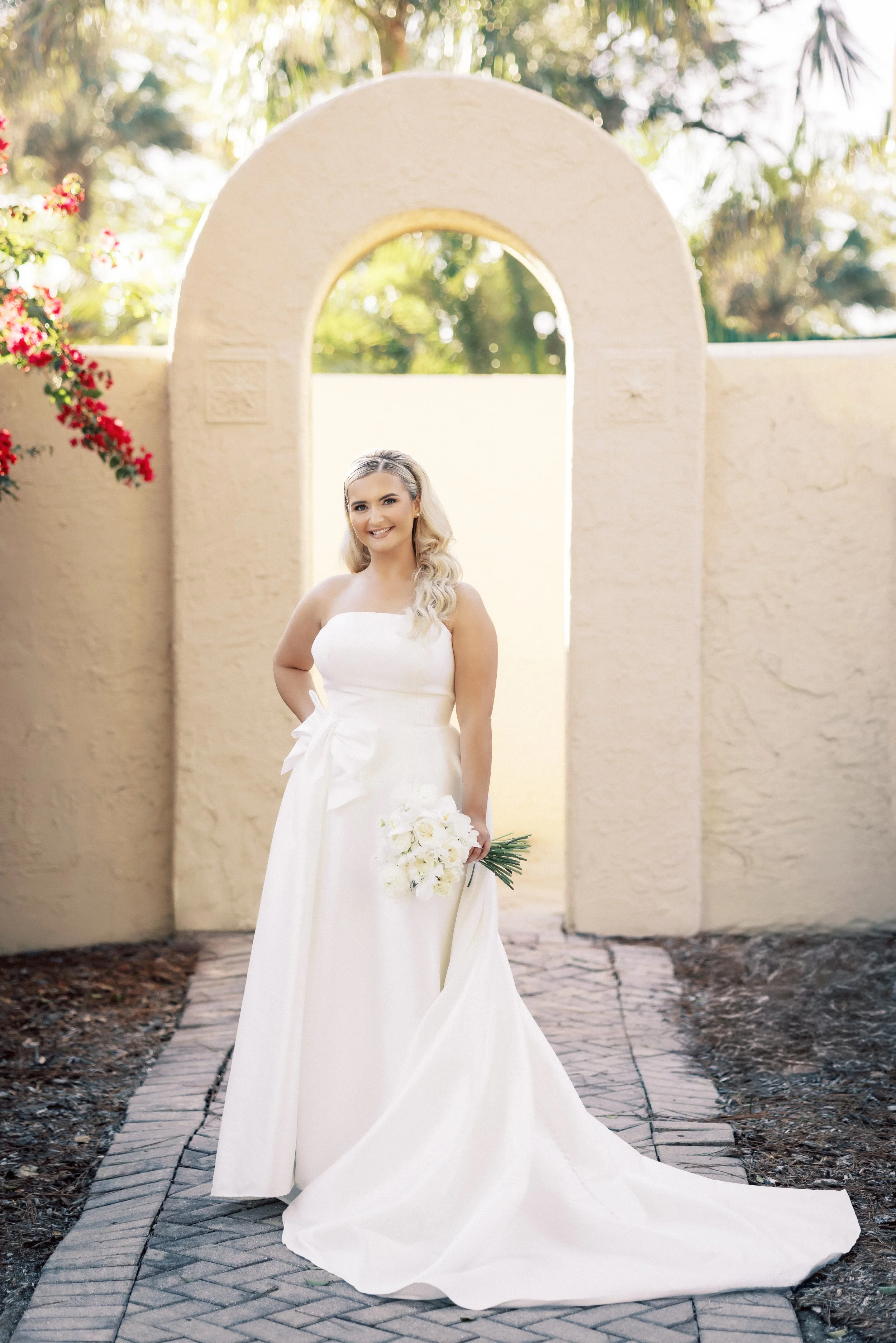A smiling bride in a white wedding dress holding a bouquet of white flowers standing in front of an arched doorway with a stucco wall and greenery in the background.