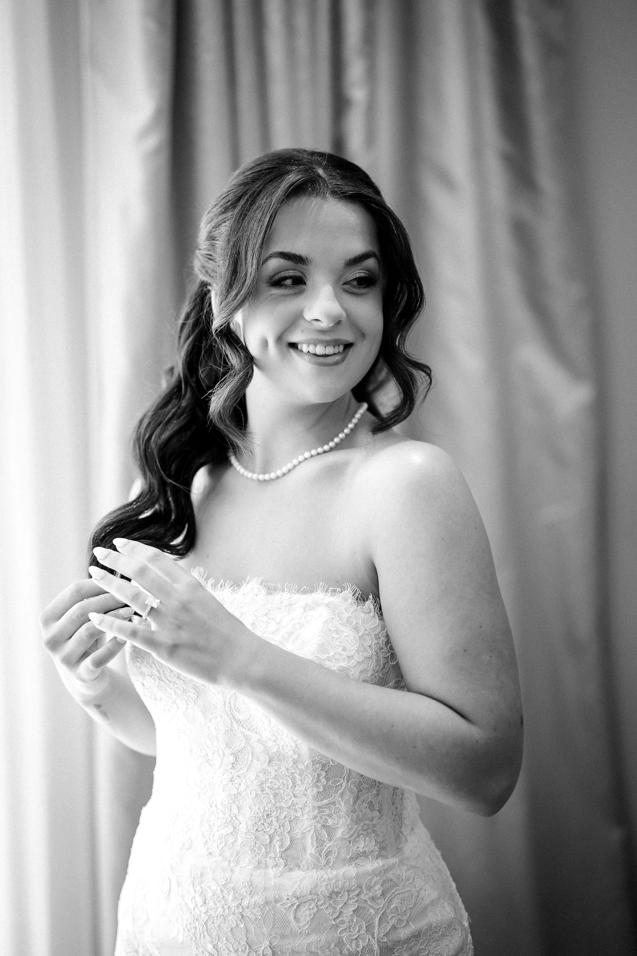 Black and white photo of a bride with wavy hair, wearing a strapless lace wedding gown, pearl necklace, and ring, smiling and looking to the side, with curtains in the background.