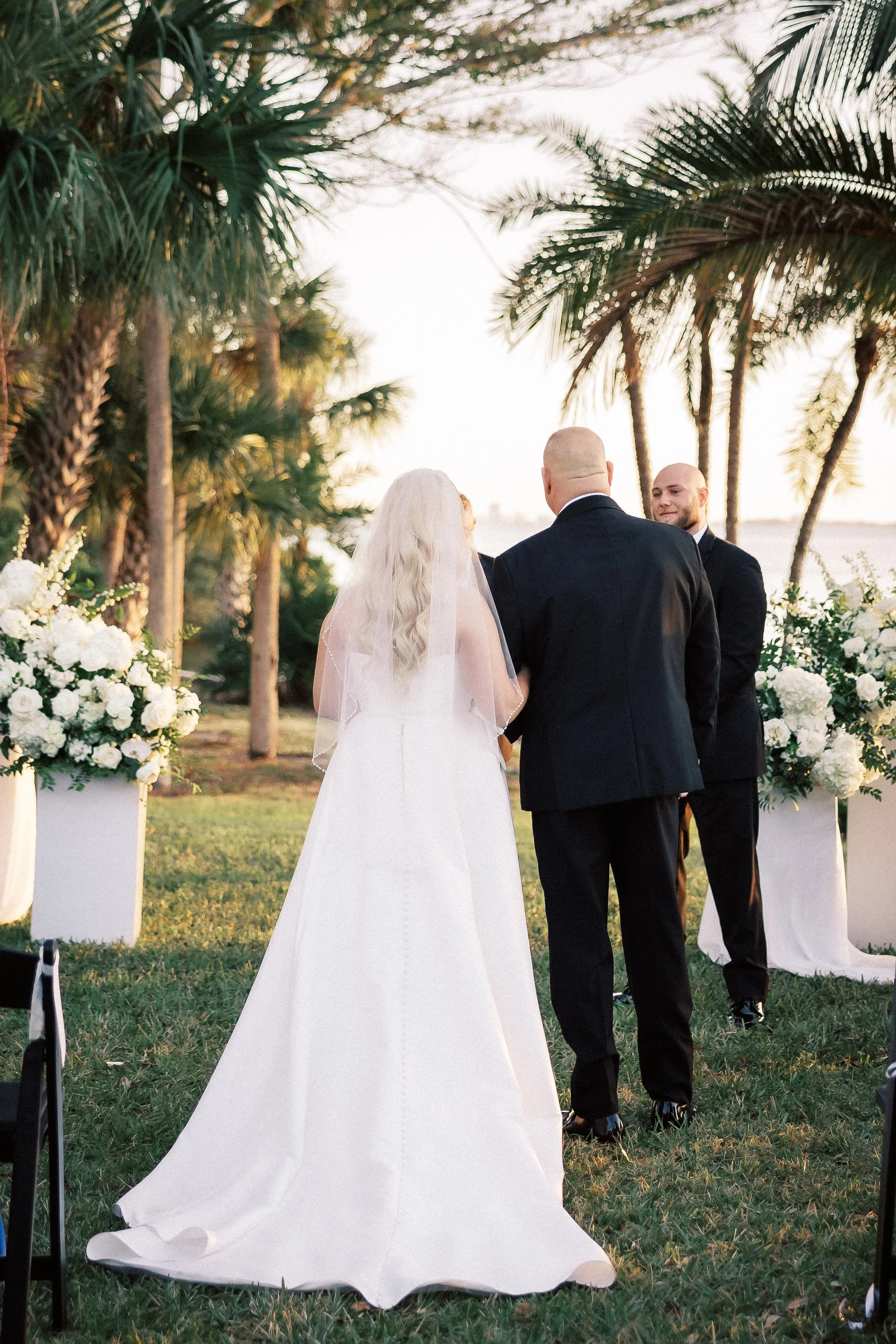 A bride with long blonde hair and a veil stands with a man, likely her father, during a wedding ceremony outdoors, with an officiant and a groom present, set against palm trees and floral arrangements.