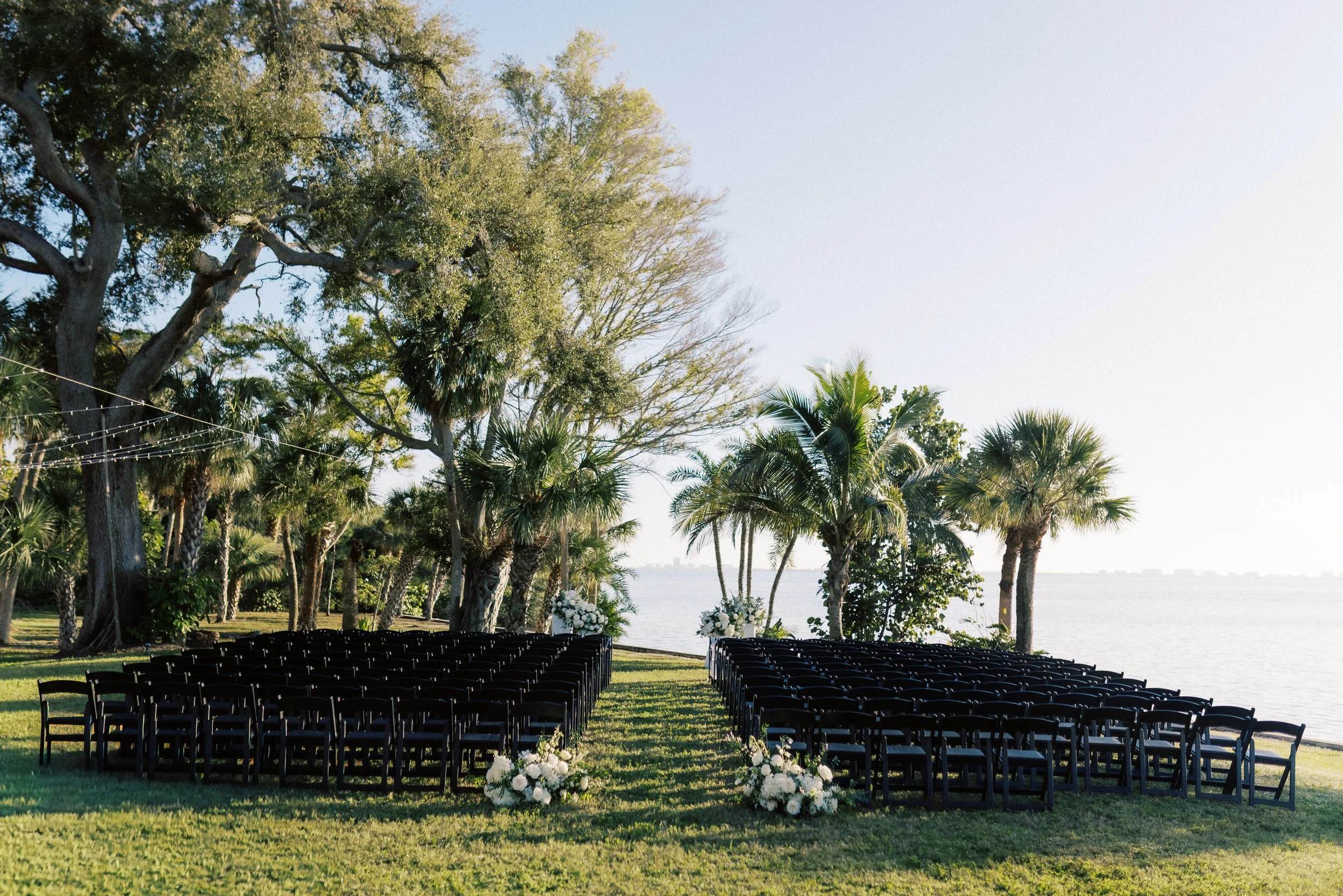 Outdoor wedding setup with rows of black chairs and floral arrangements, on a lawn near a body of water with trees and palm trees in the background.