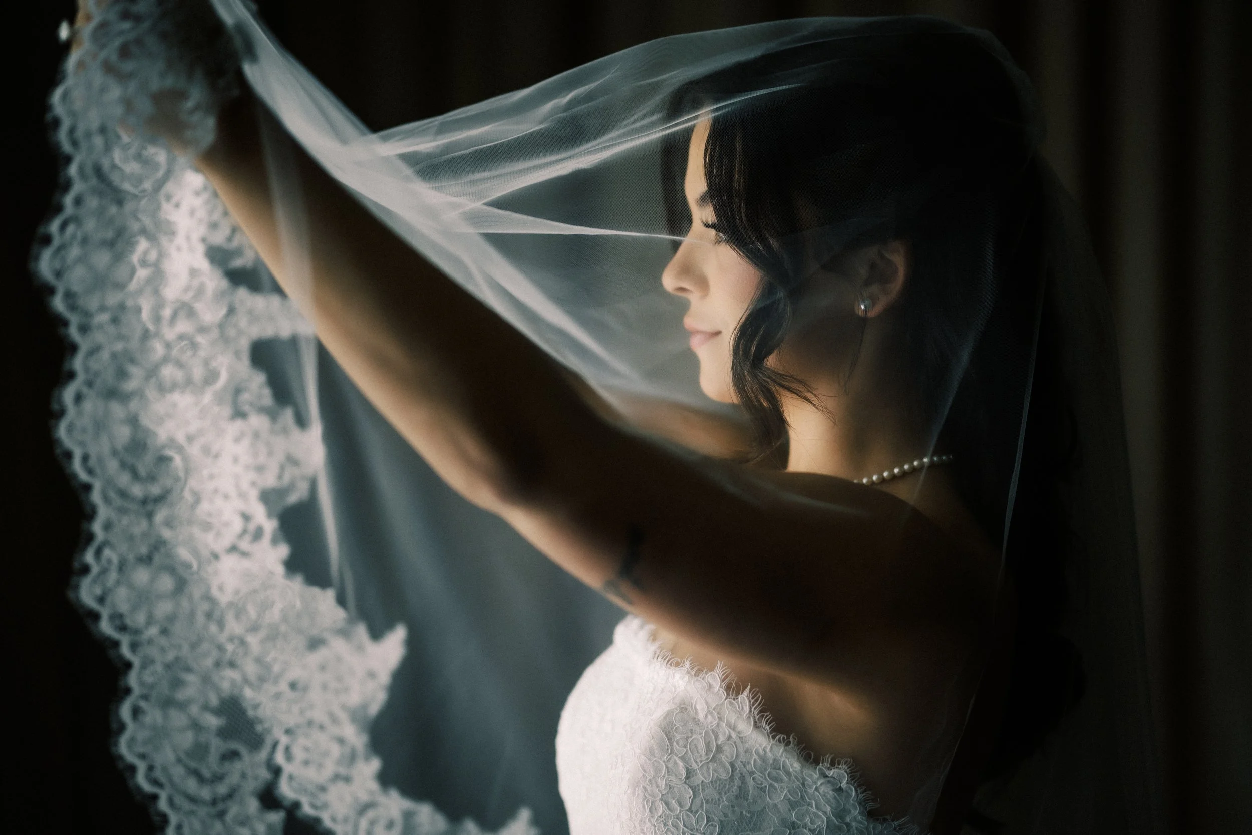 A bride with dark hair and pearl earrings, smiling softly, is lifting her wedding veil. She is wearing a white lace wedding dress with a pearl necklace, posed in profile against a dark background.