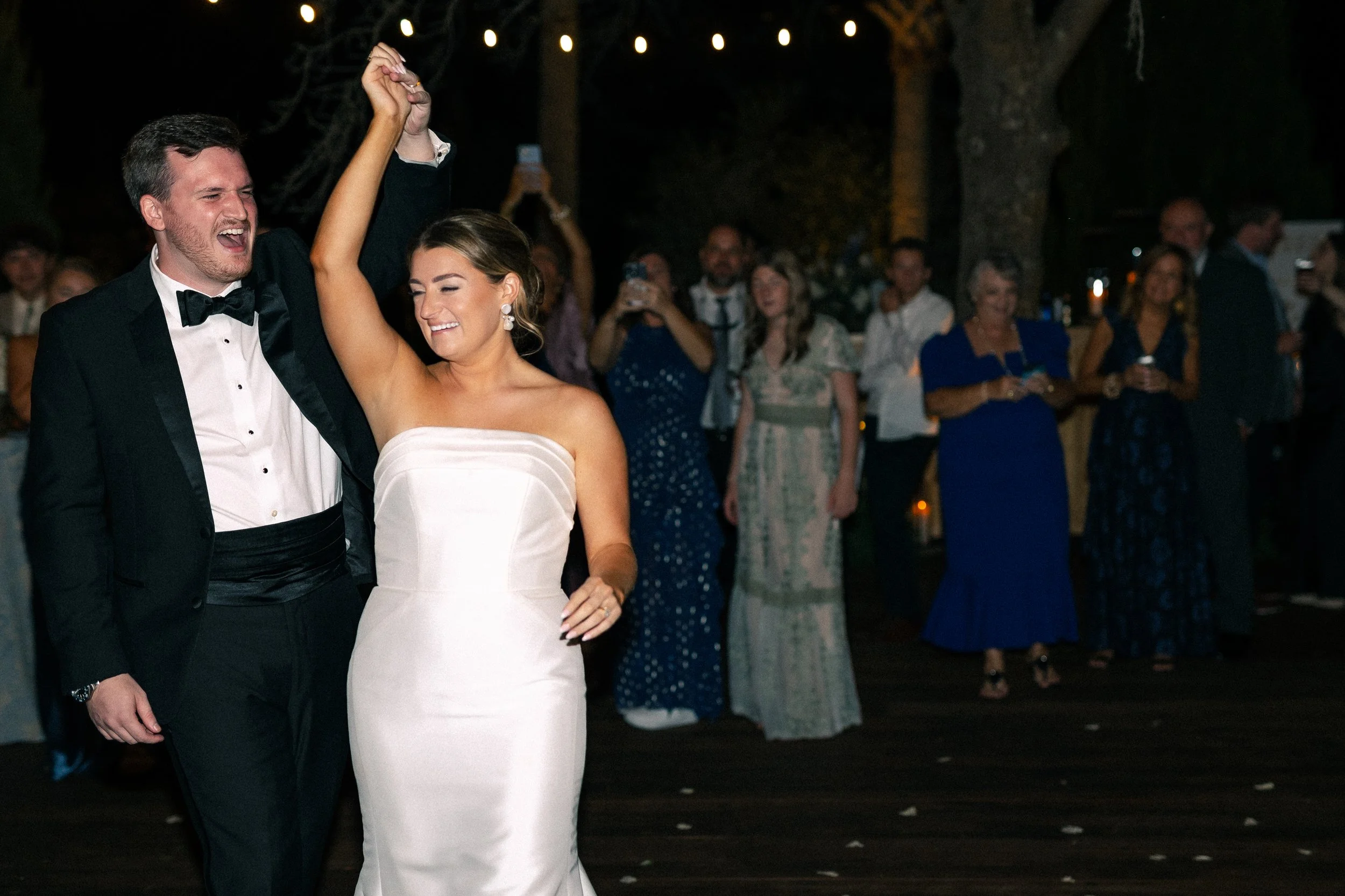 A bride and groom dancing at their wedding reception, surrounded by friends and family outdoors at night.