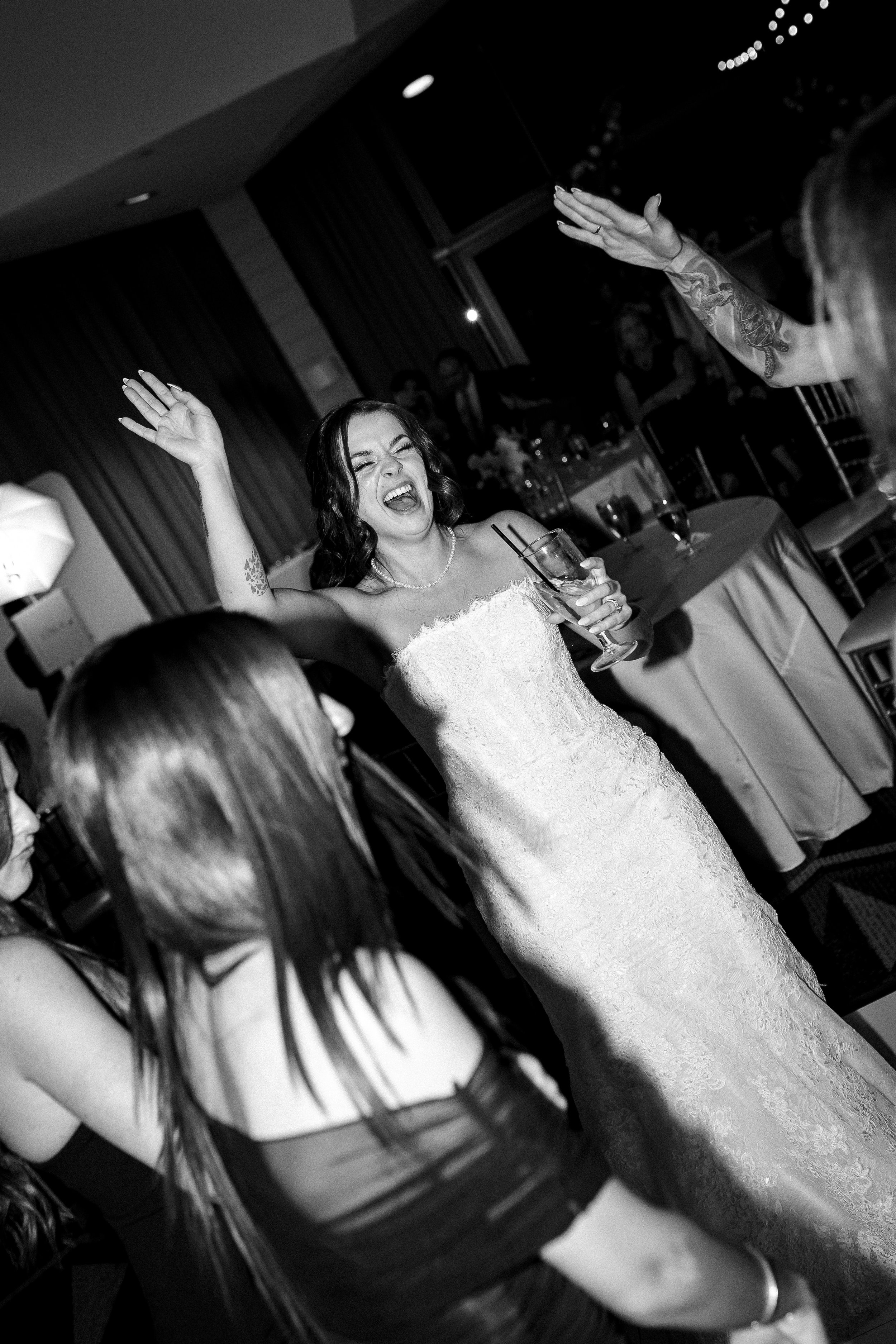 A group of women dancing and celebrating at a wedding reception, with a bride in a white lace wedding dress holding a drink, smiling and raising her hand.