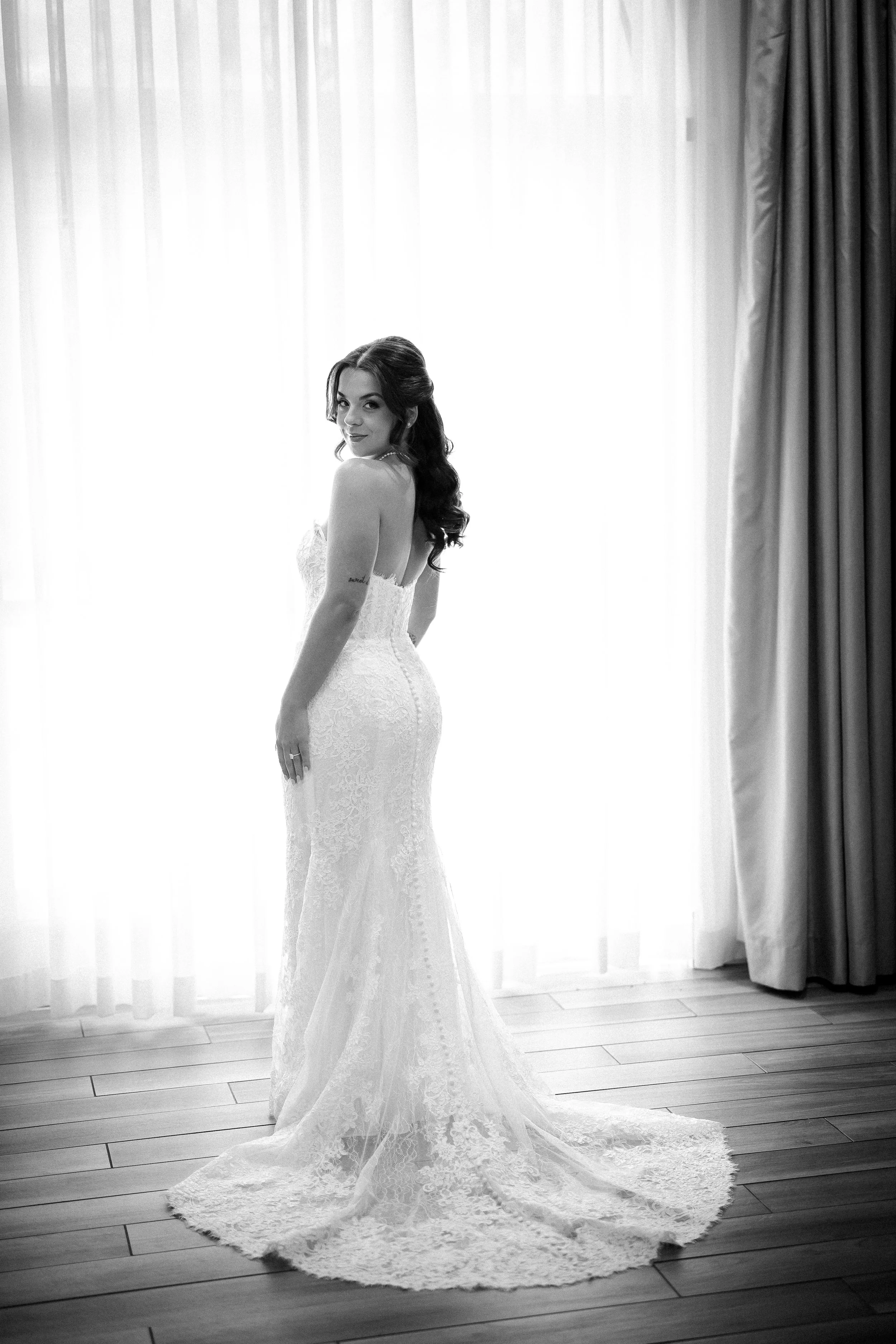 A woman in a lace wedding gown posing in front of a sheer curtain and drapes, with a wooden floor beneath her.