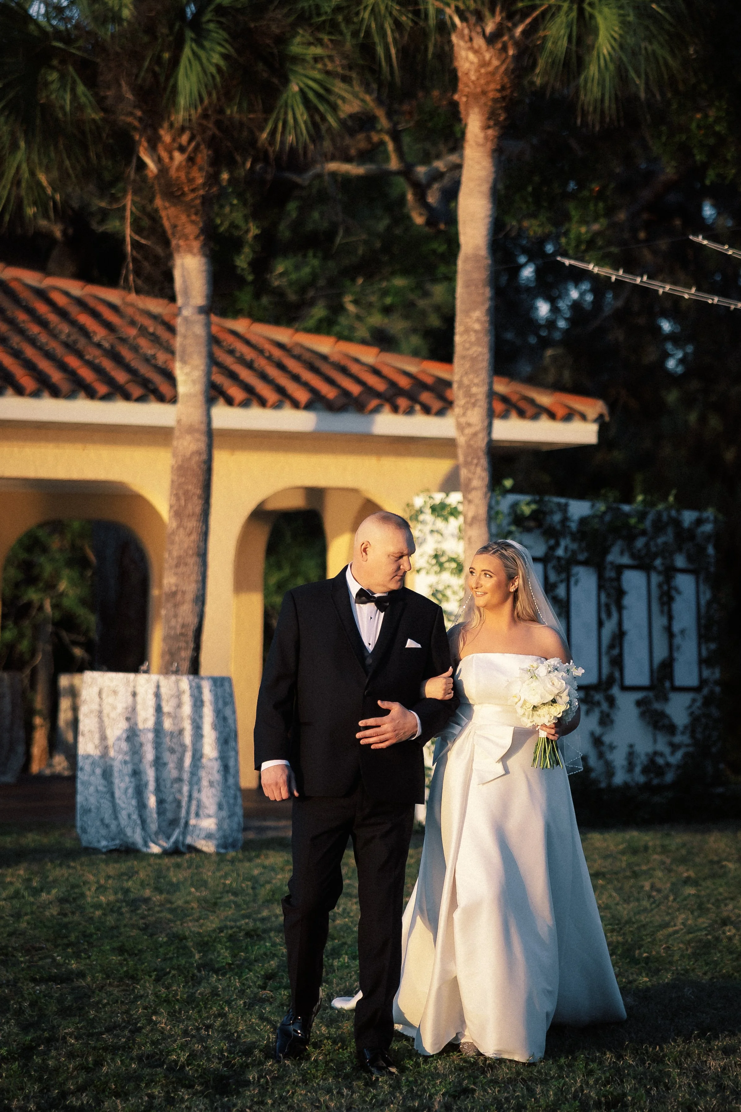 A bride in a white wedding gown holding a bouquet walking with a groom in a black tuxedo at an outdoor wedding venue with palm trees.