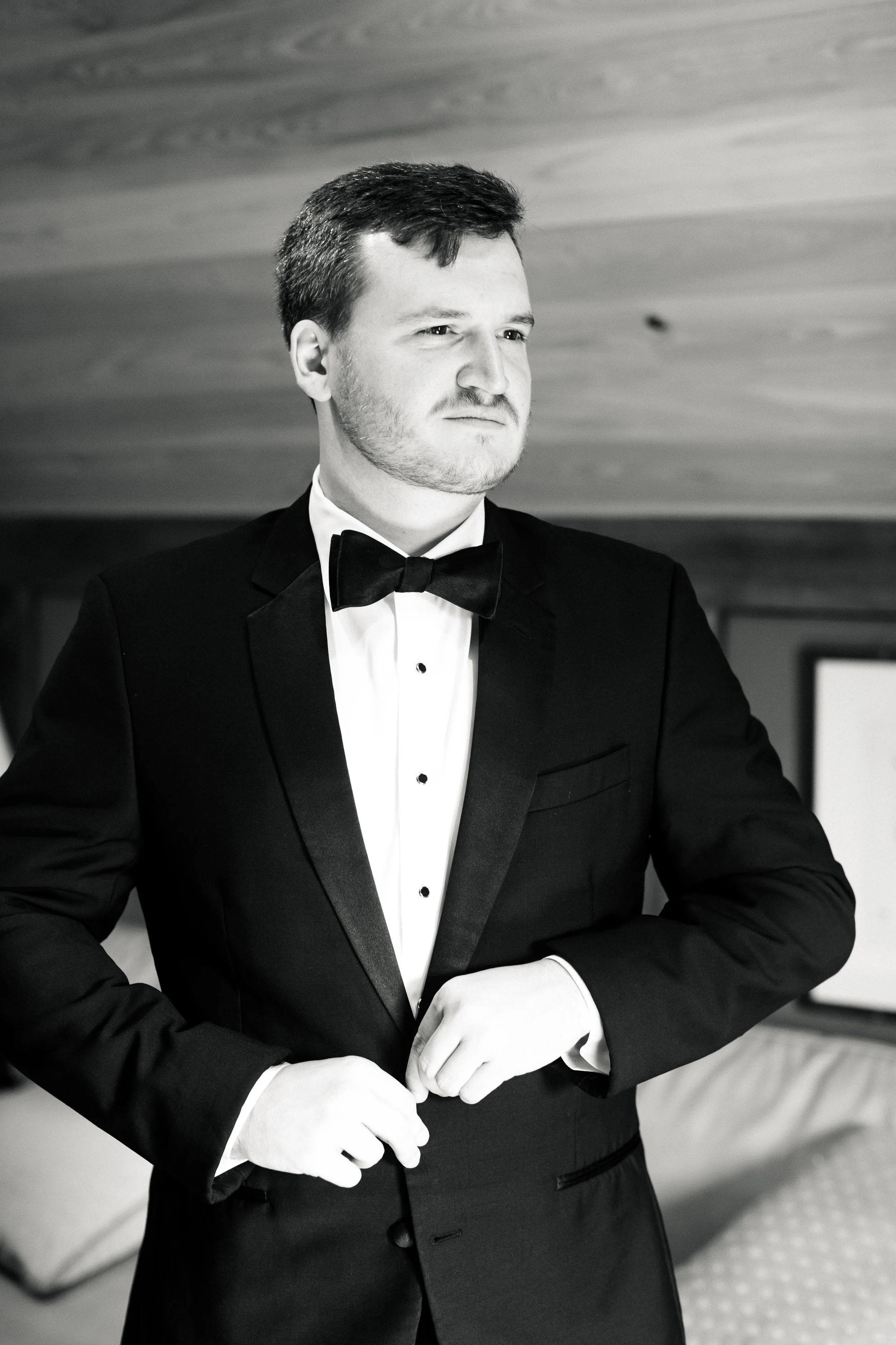 A man dressed in a tuxedo, adjusting his jacket, with a serious expression, standing indoors.