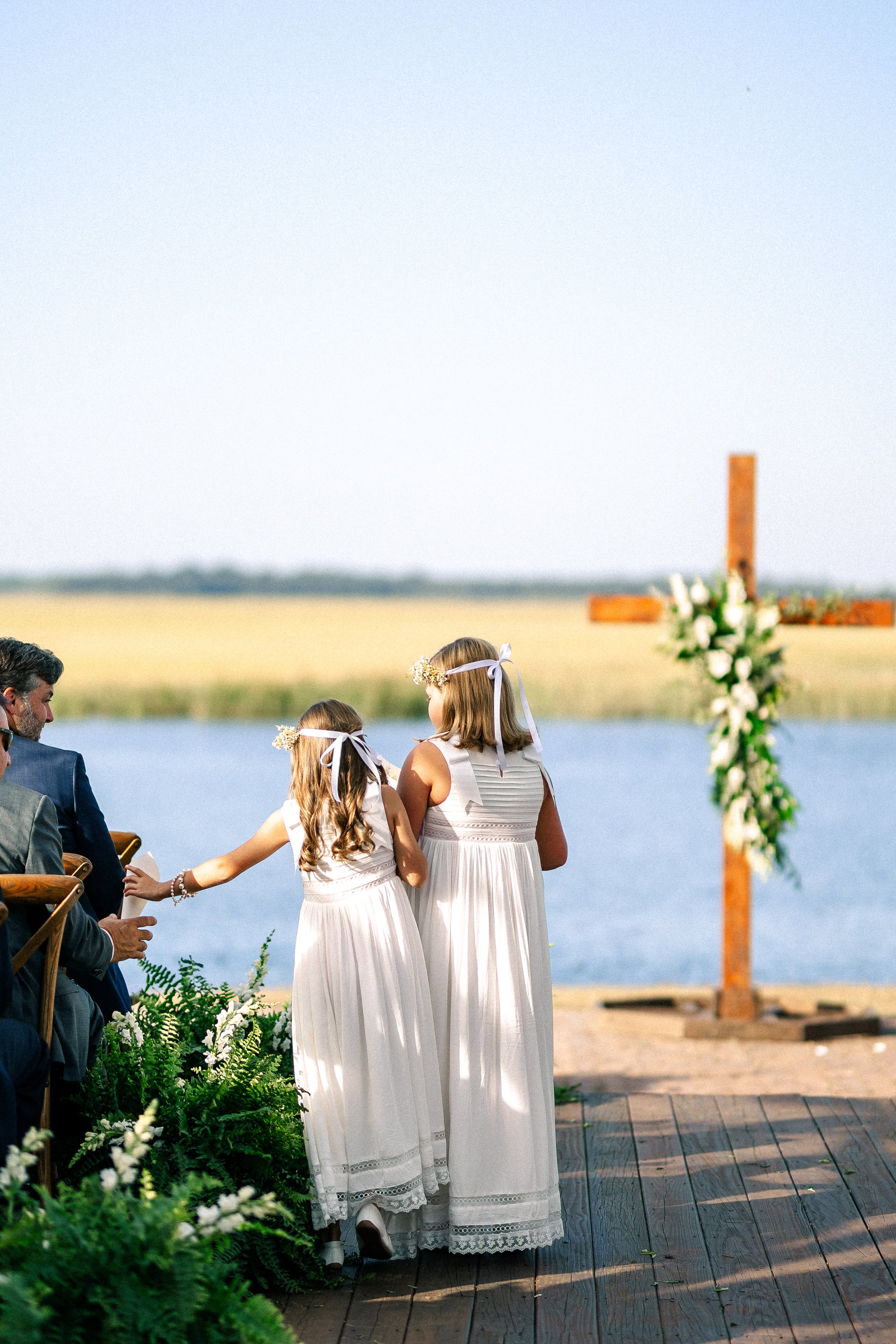 A wedding ceremony outdoors near a body of water with two young girls dressed in white, standing in front of a wooden cross decorated with flowers, holding hands.