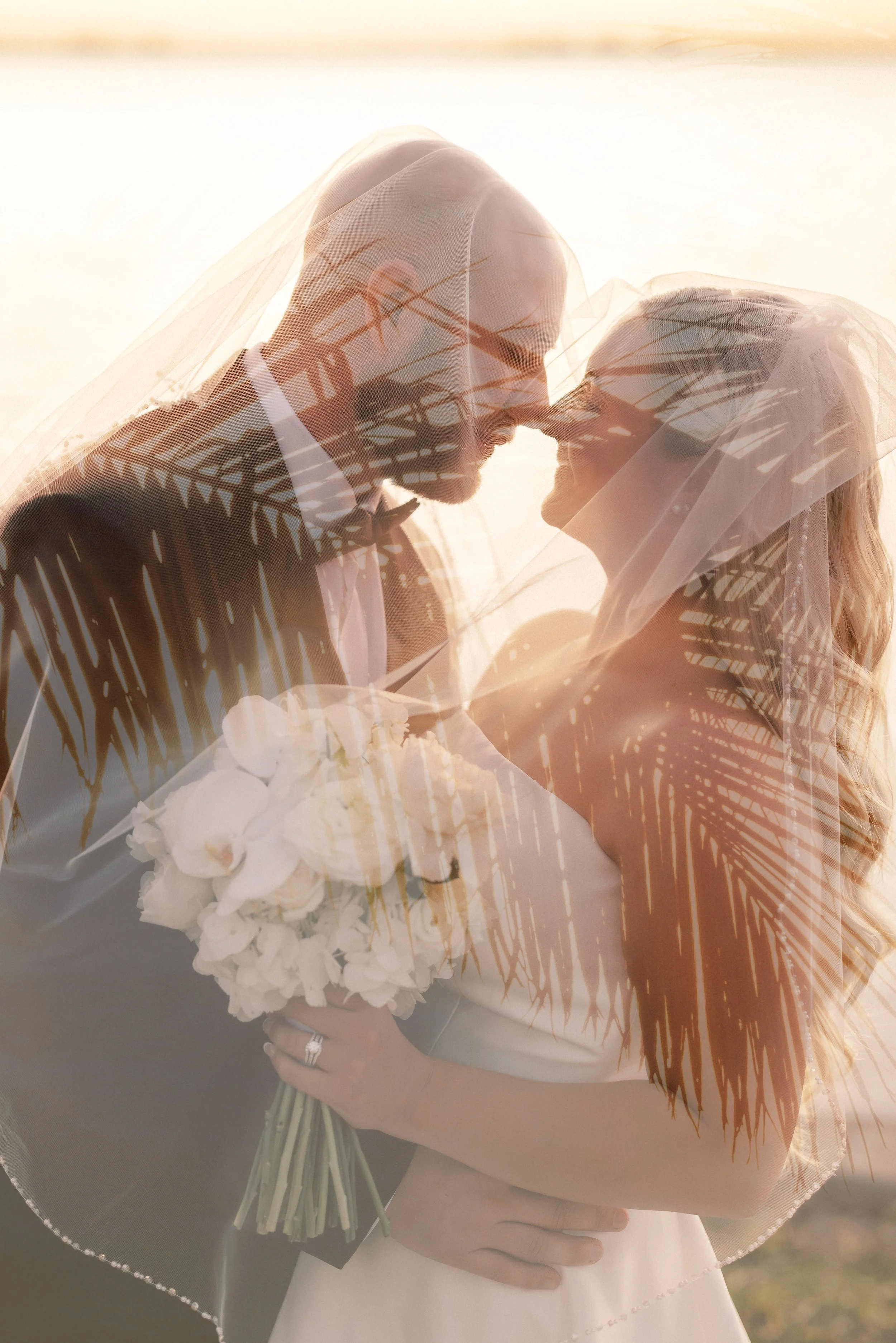 A bride and groom under a wedding veil, holding a bouquet of white flowers, sharing a moment at sunset with palm leaf reflections overlayed.