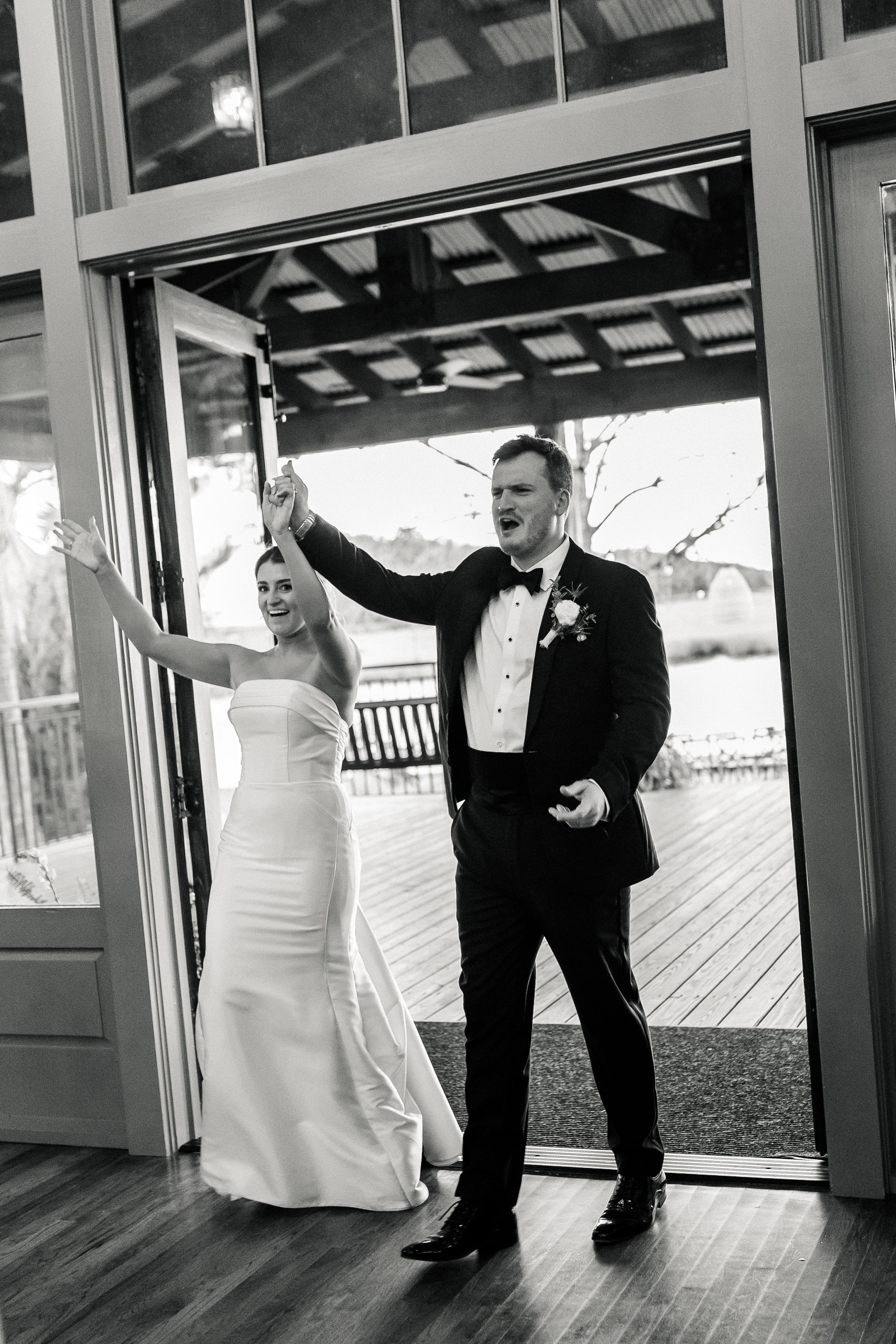 Black and white photo of a bride and groom entering a room. The bride is wearing a strapless wedding gown and smiling with her arms raised. The groom is wearing a tuxedo with a bow tie, making an expressive gesture.
