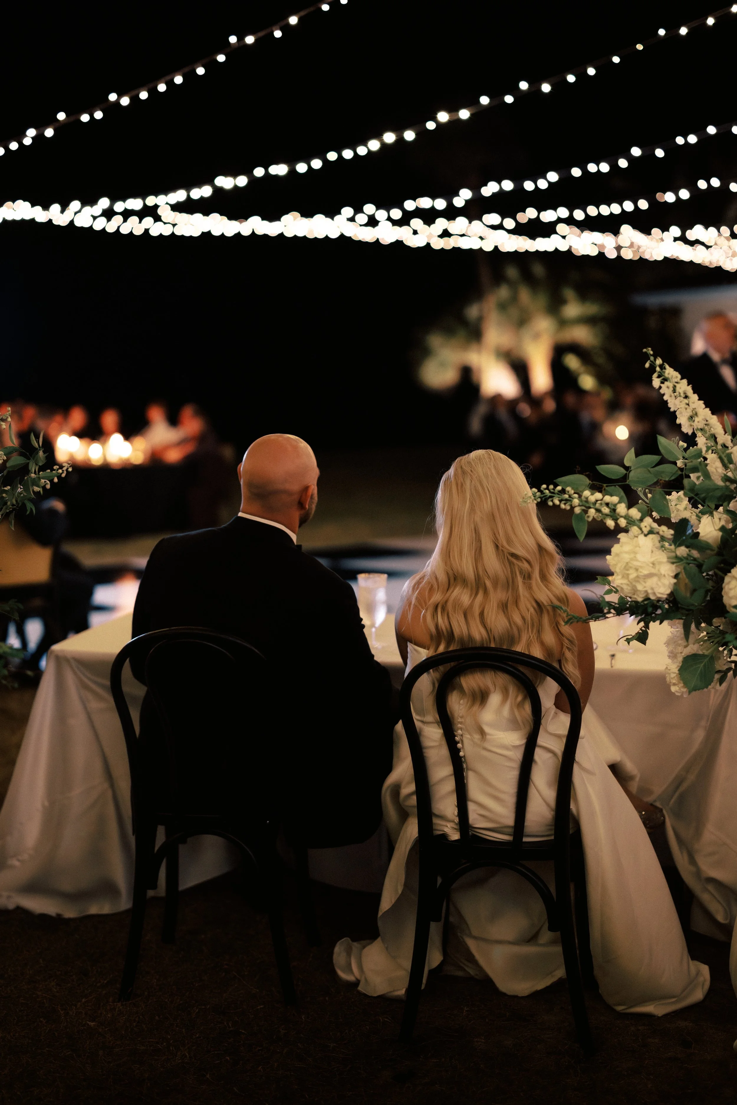 A bride and groom sitting at a wedding reception under string lights at night, with floral arrangements on the table.
