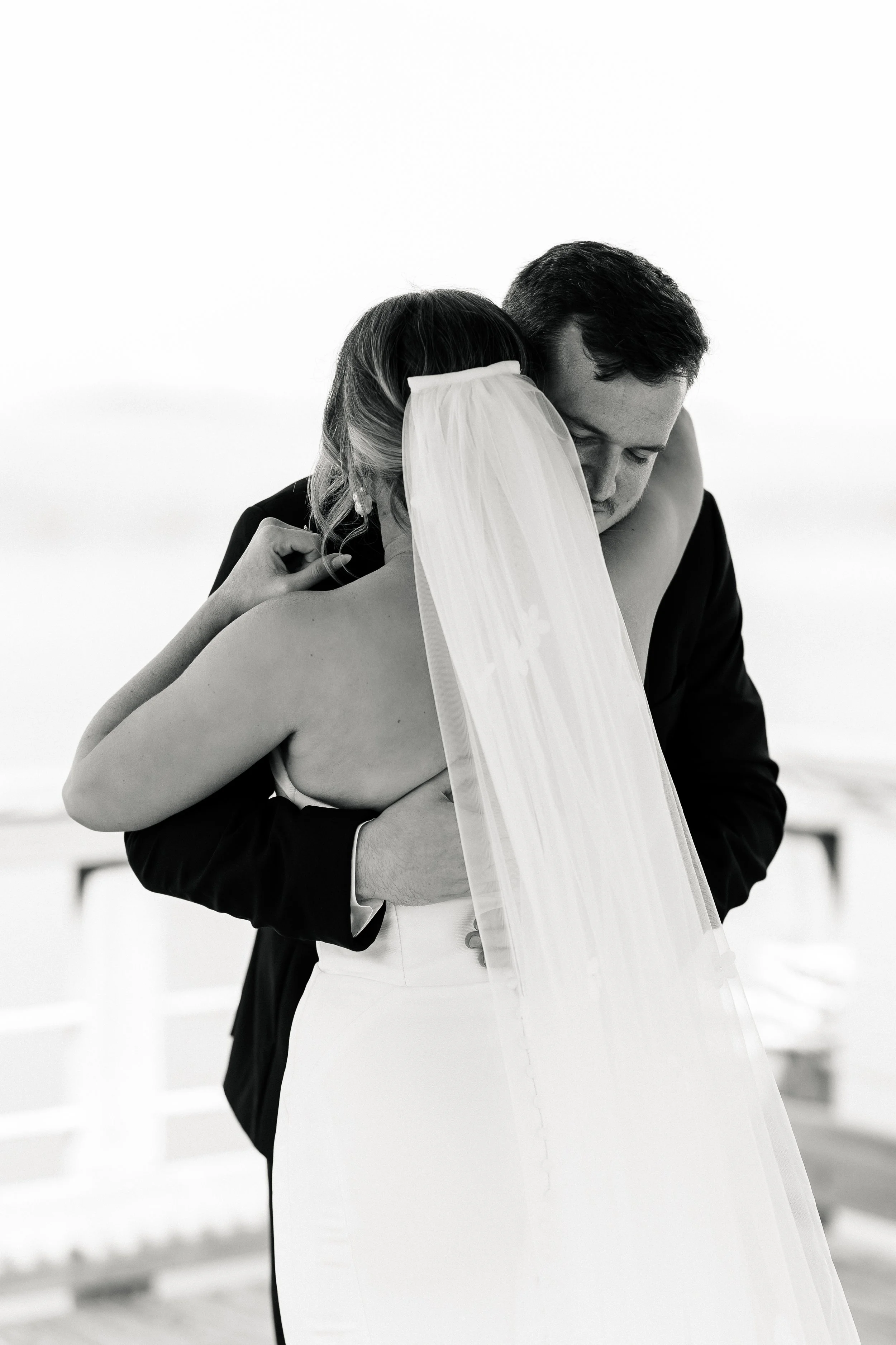 A black and white photograph of a bride and groom embracing each other. The bride is wearing a veil and a strapless dress, while the groom is dressed in a suit.