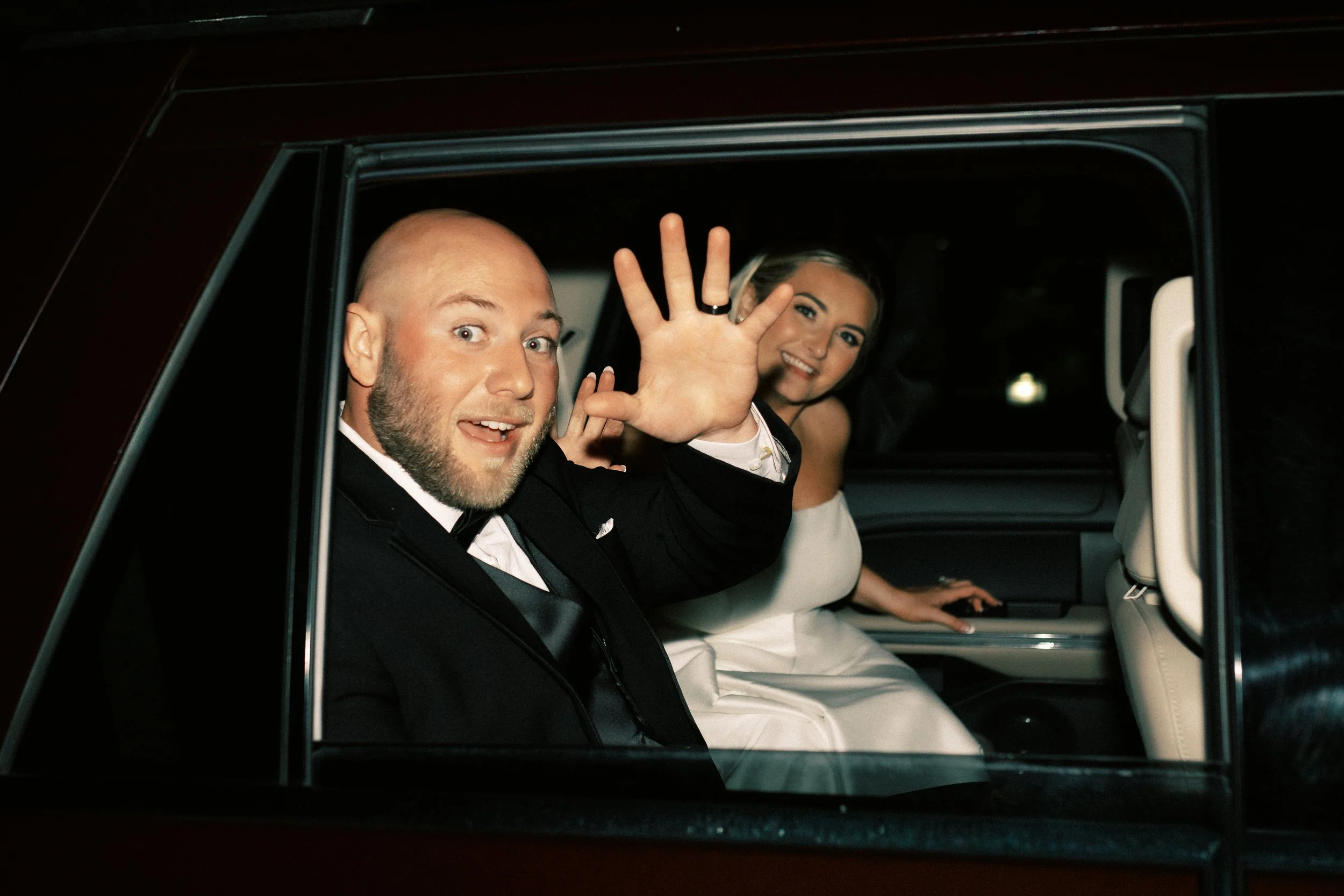 A smiling man in a tuxedo waving from the window of a limousine, with a woman in a white dress inside the vehicle.