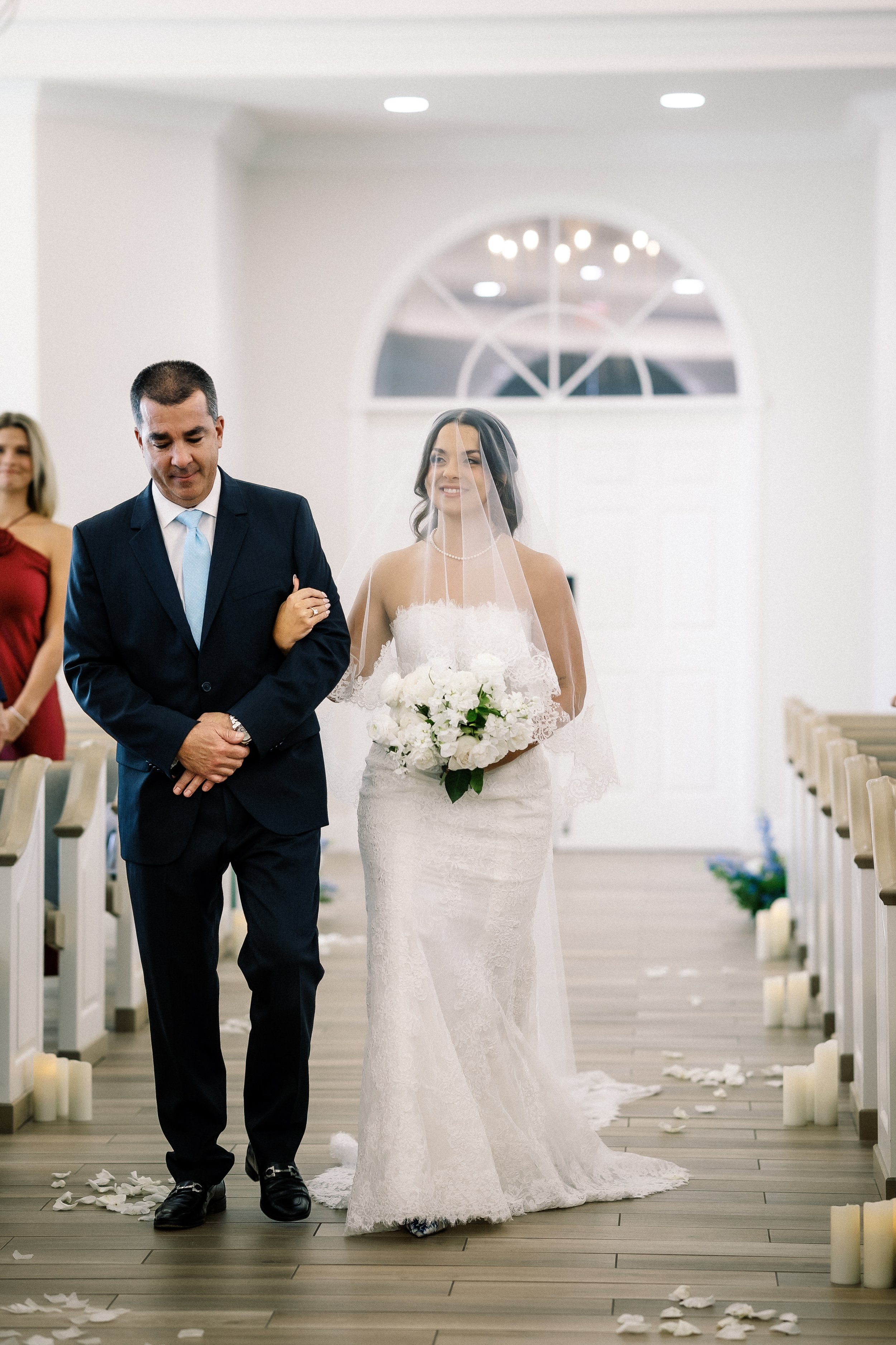 Bride walking down the aisle with her father during a wedding ceremony in a bright indoor chapel.