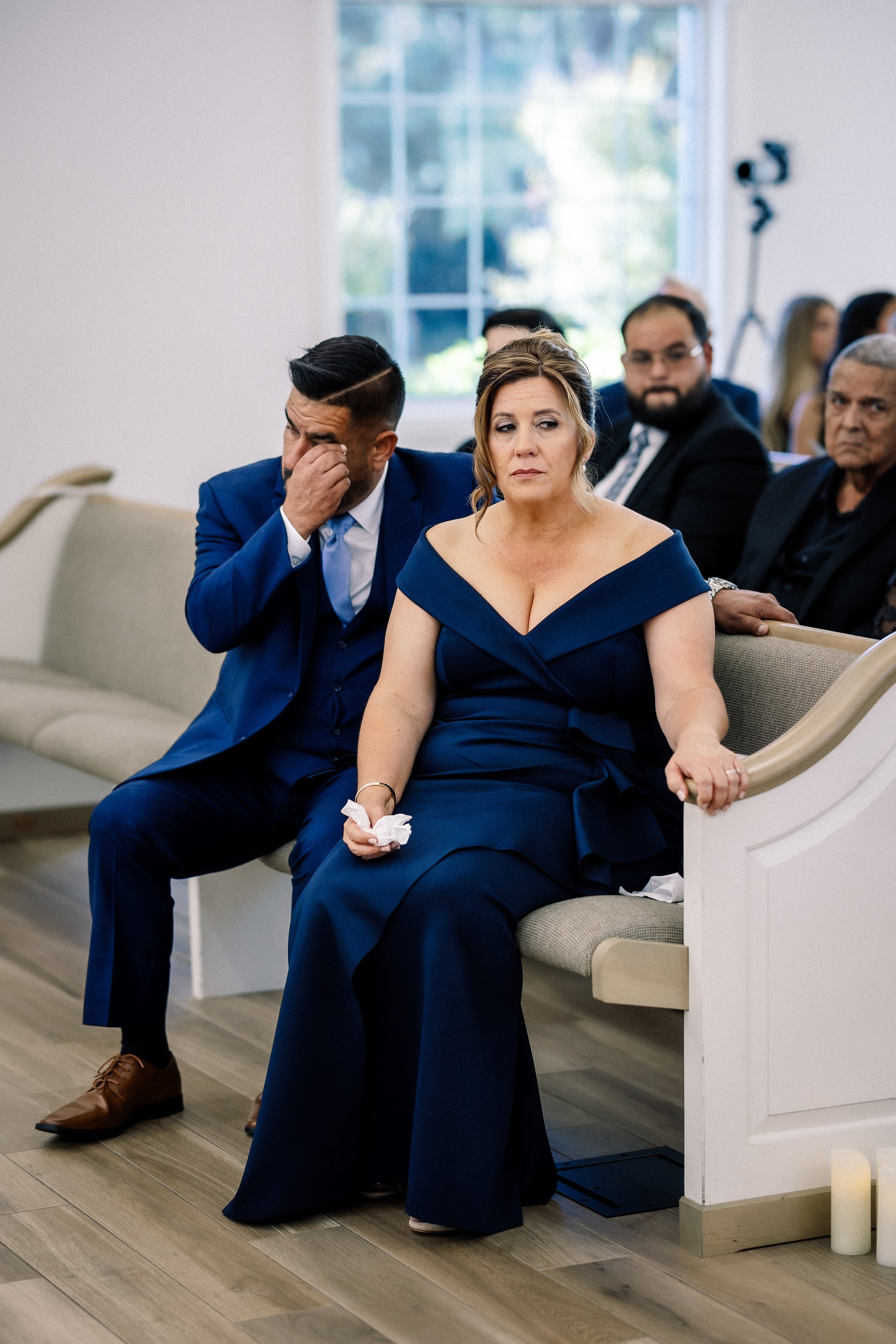 People sitting in pews at a formal event, with one woman in a navy blue gown looking upset and a man beside her wiping his eyes.