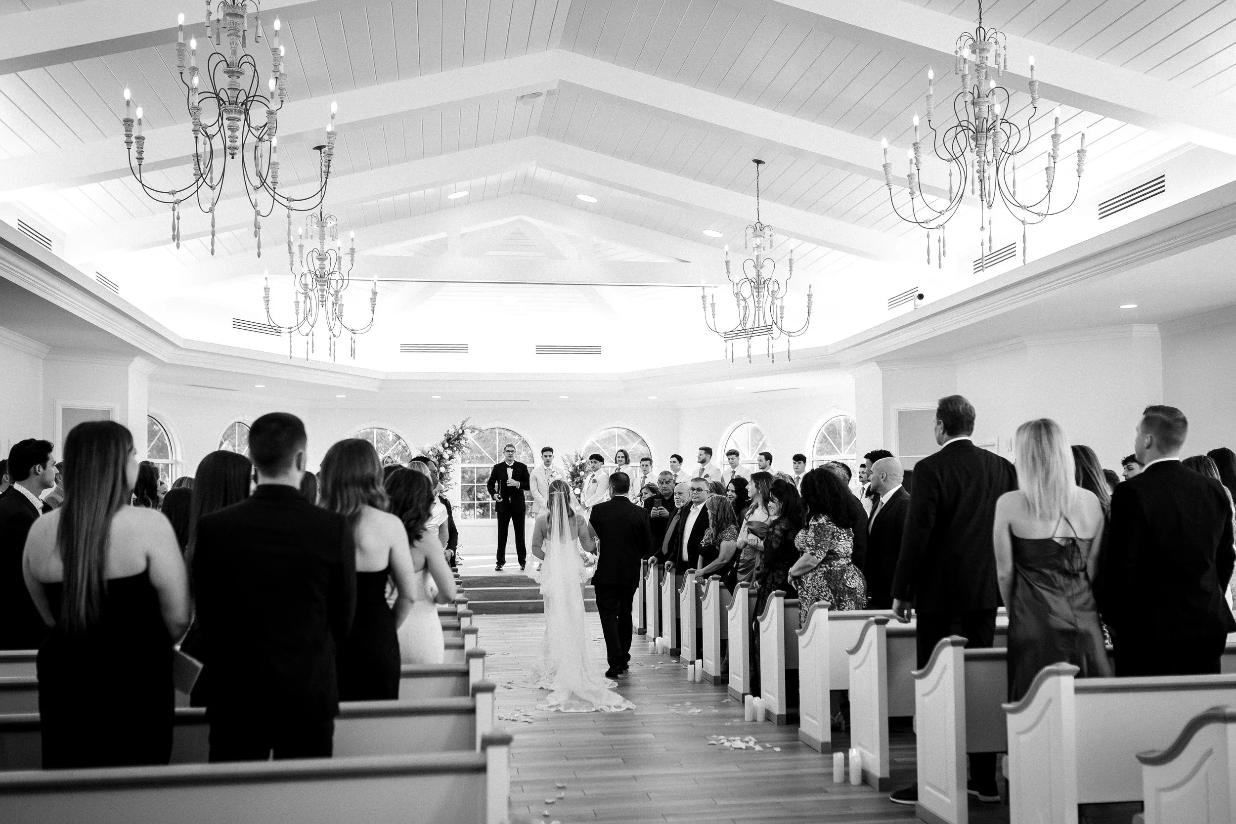 Black and white photo of a wedding ceremony inside a spacious, well-lit hall with high ceilings and chandeliers. The bride and groom walk down the aisle together as guests stand and watch on either side.