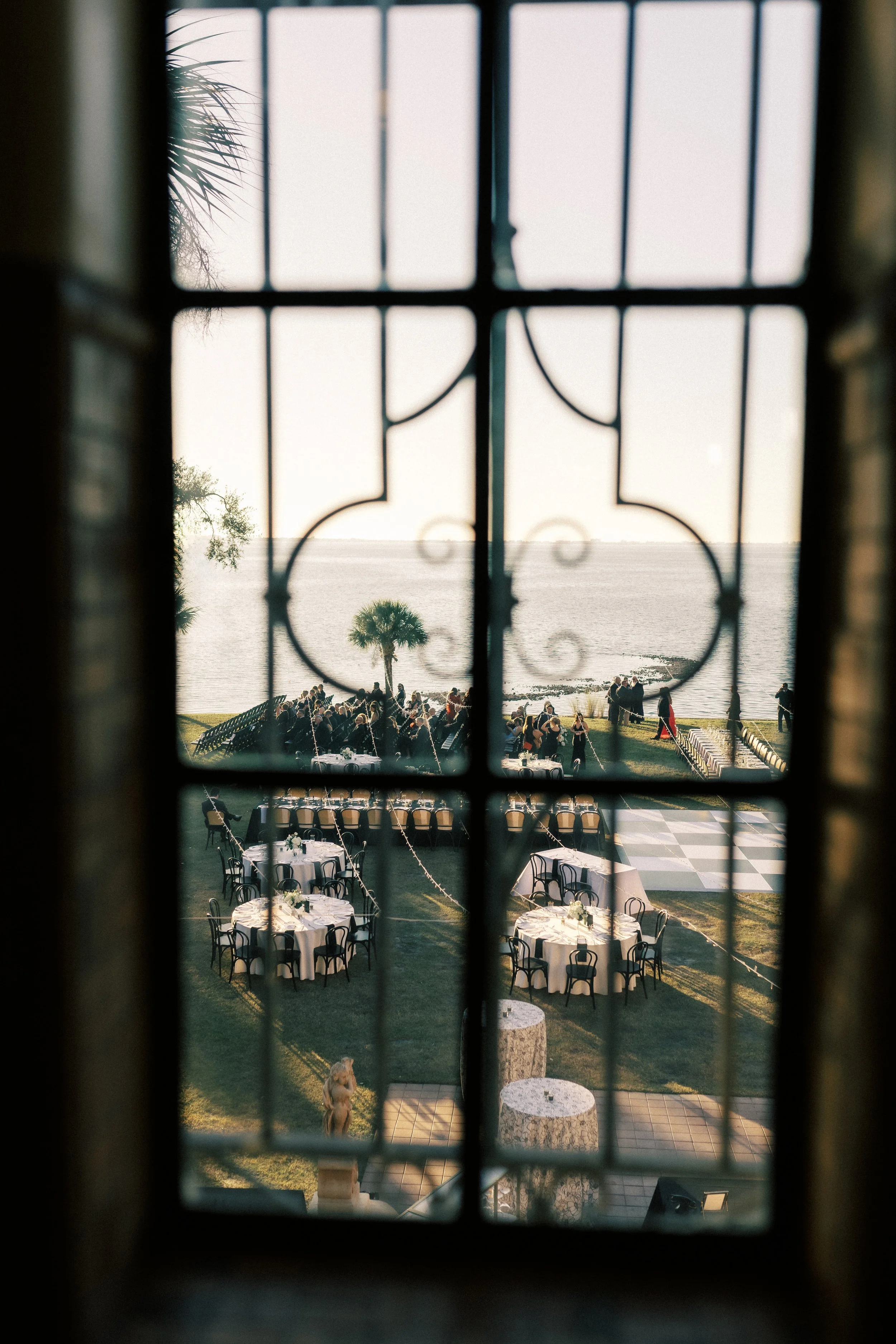 A view of an outdoor event by the water, seen through a window with decorative iron bars. The scene includes tables with white tablecloths, chairs, and people gathered near the shoreline, with the ocean and a few palm trees in the background.