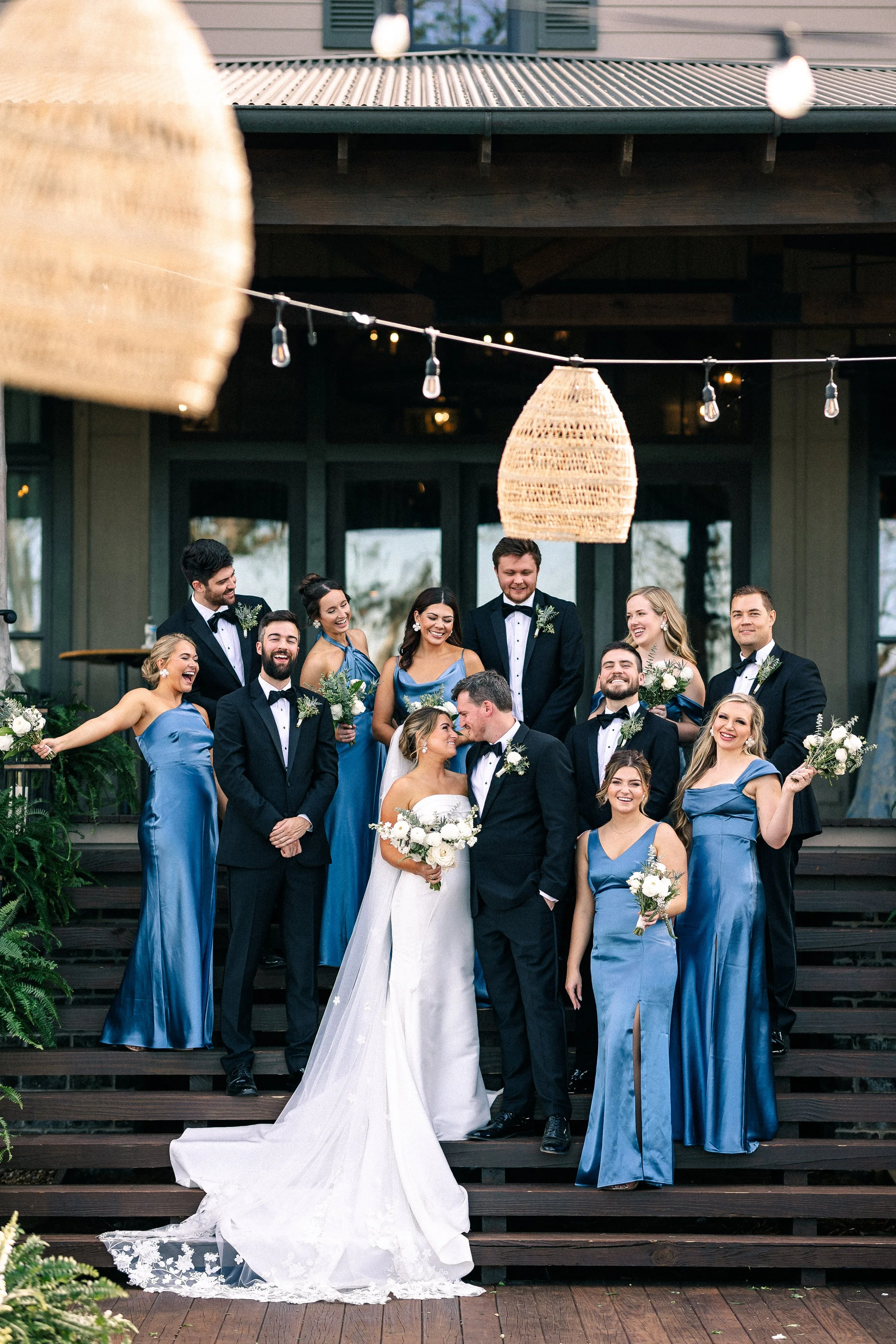Wedding party posing outdoors on steps at a rustic venue, with the bride and groom in the center, surrounded by bridesmaids in blue dresses and groomsmen in tuxedos."}