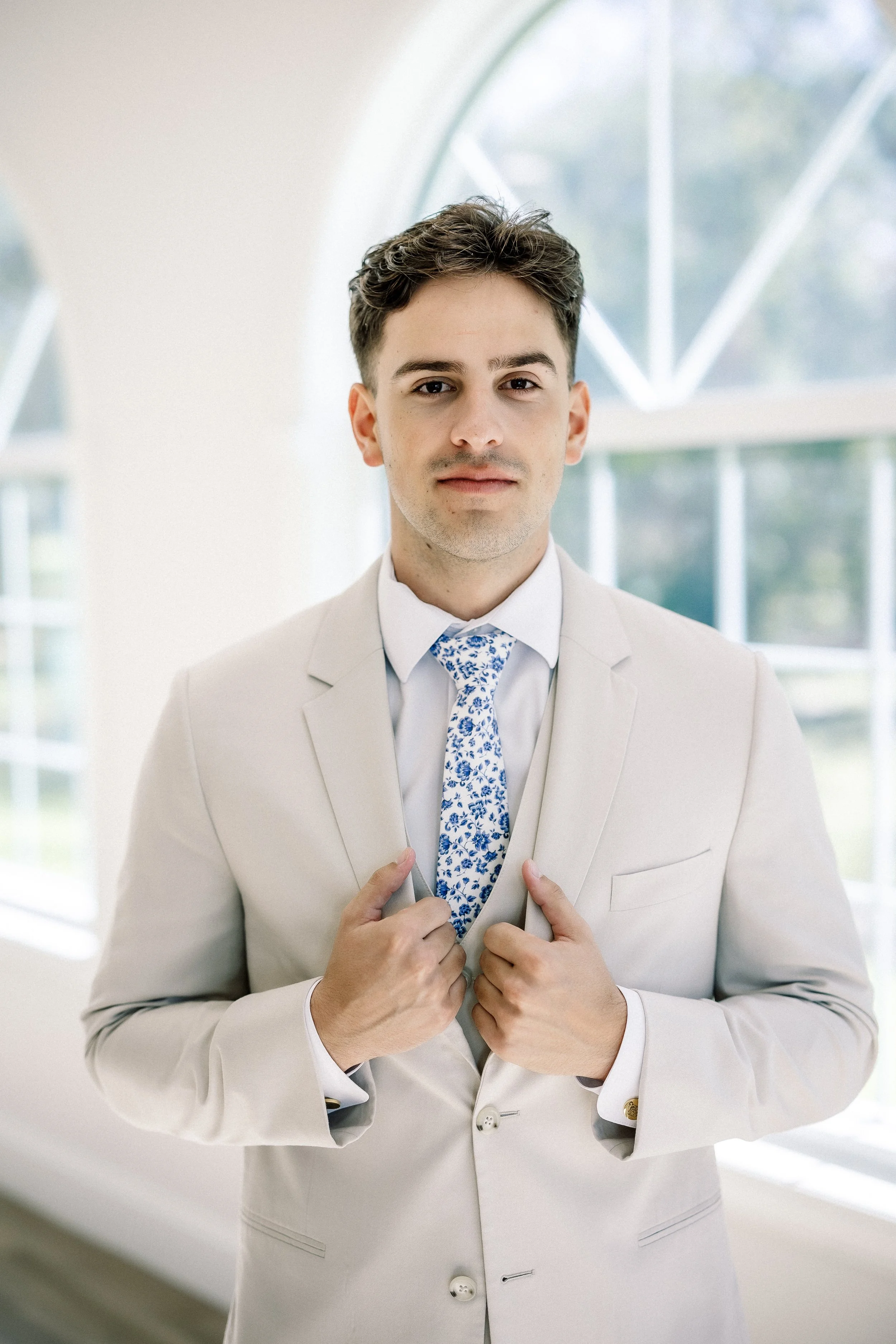 A young man in a light-colored suit and a floral tie, standing indoors in front of large windows, looking confidently at the camera while holding his jacket lapels.