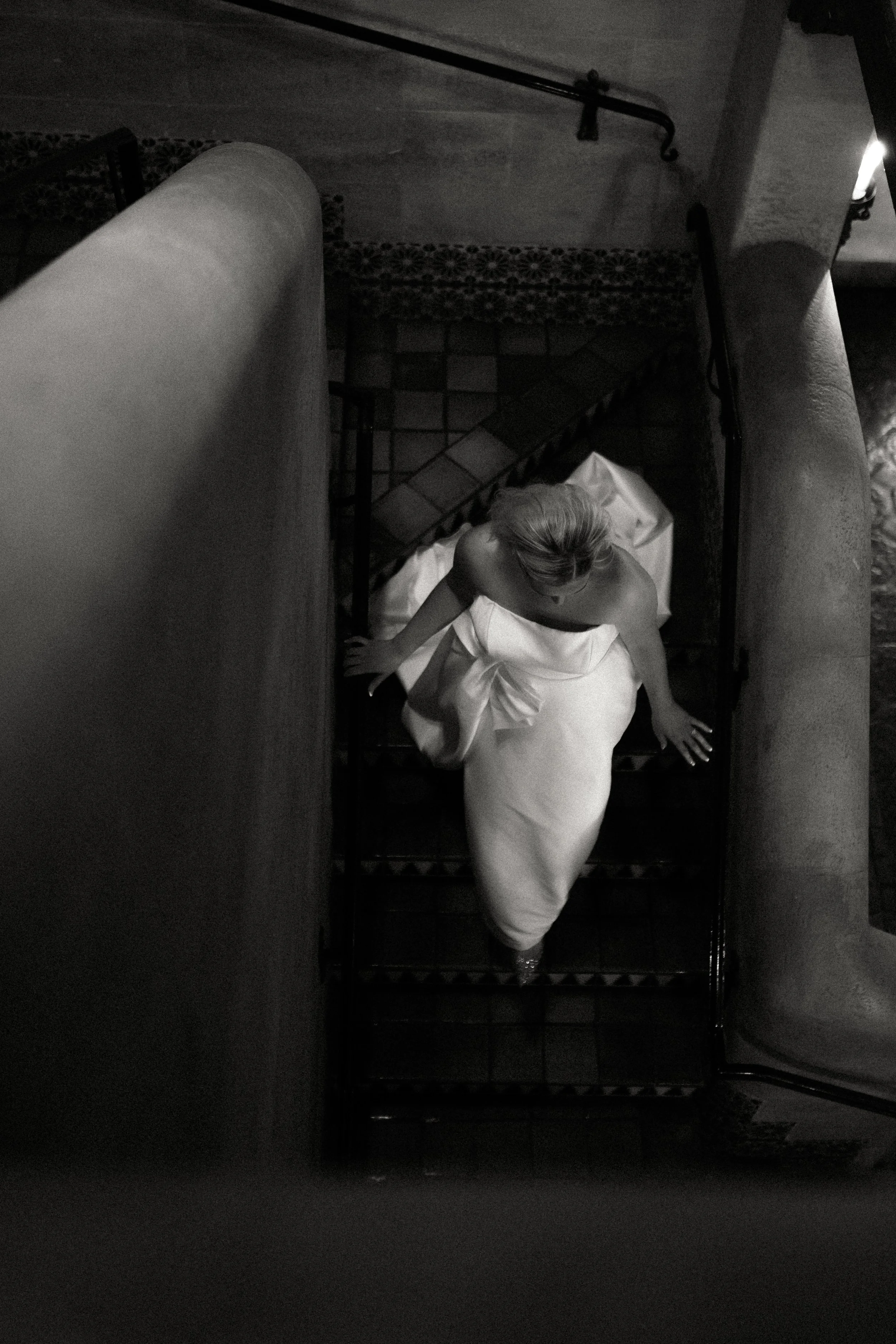 A woman in a white dress descending a staircase viewed from above. The staircase has tile steps and metal handrails.