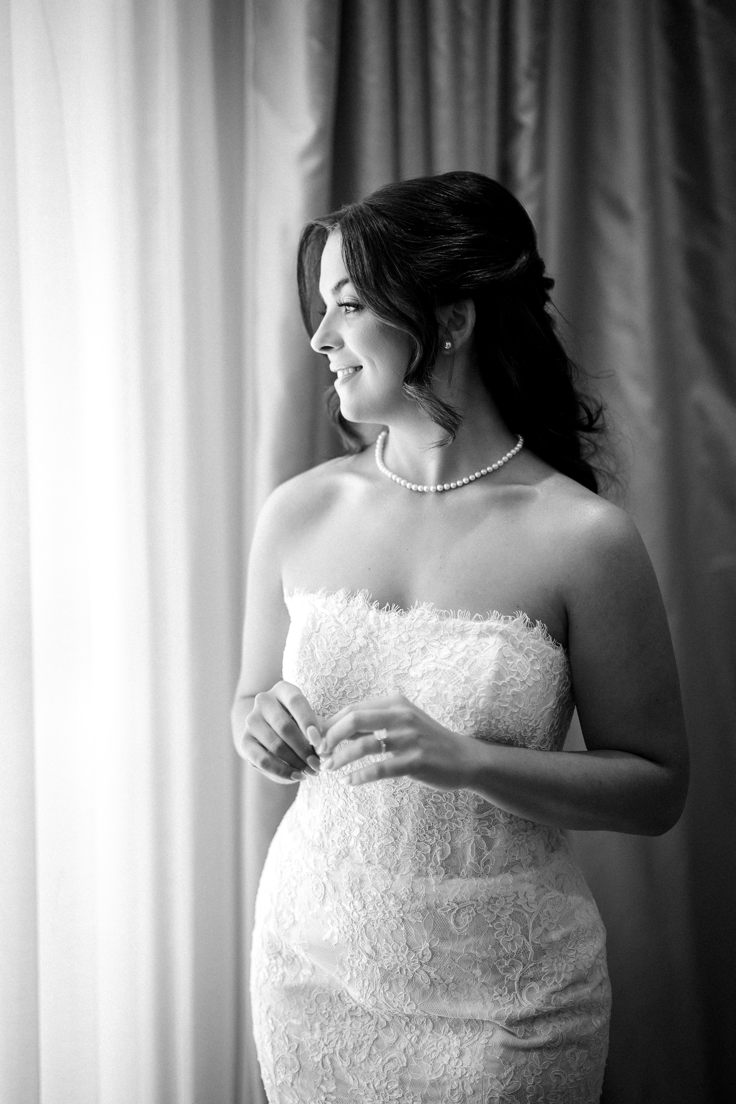 A bride in a strapless lace wedding dress, pearl necklace, and earrings, standing near a window with curtains, smiling and looking to the side.